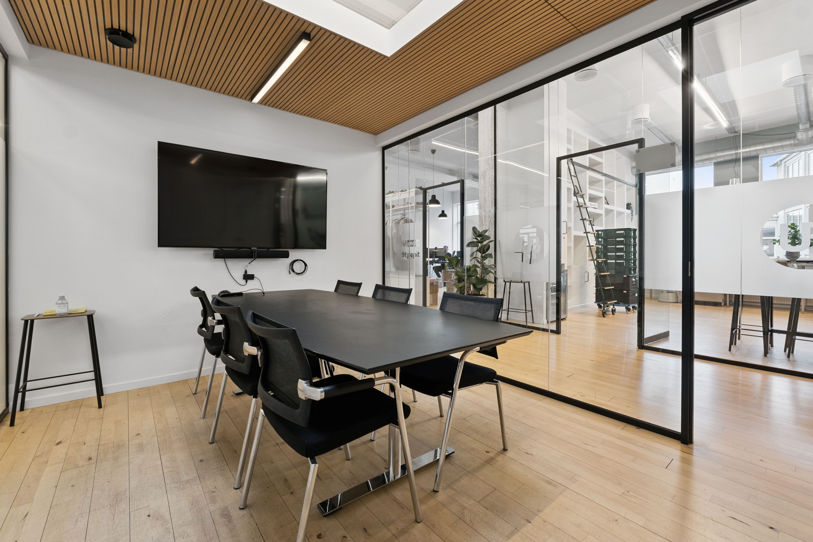 Moderne konferencelokale med lyse trægulve, glasvægge, højloft og mørk konferencebord og stole til 12 personer med tv og plante i enden