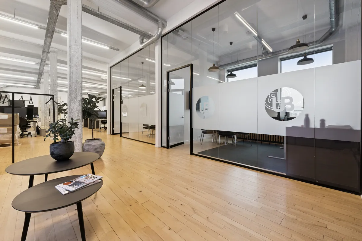 Glassvægge og lyse lofter, kontoret med moderne møbler og smukke plantepotter. To runde borde med magasiner, og flere kontorer med borde og stole.