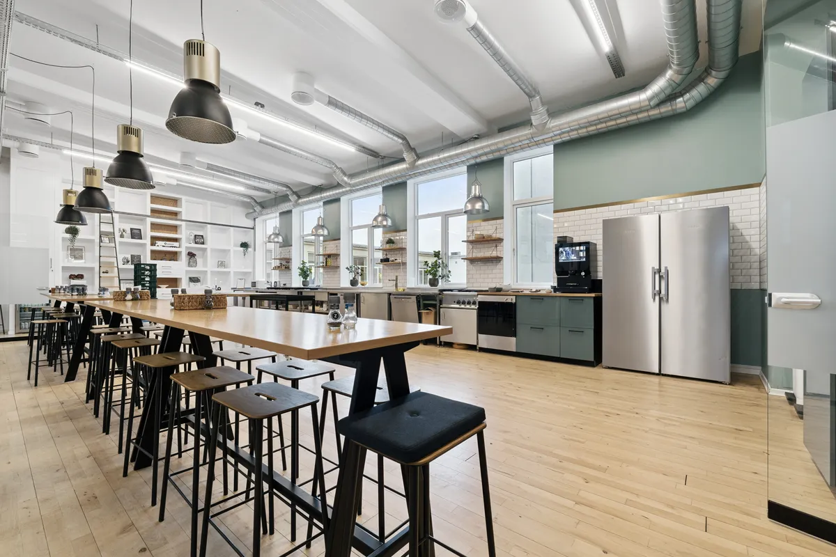 Kontorlokaler med åben køkkendel, lyse loftslamper og trægulv samt møbler og udstyr til rådighed i et industrielt miljø