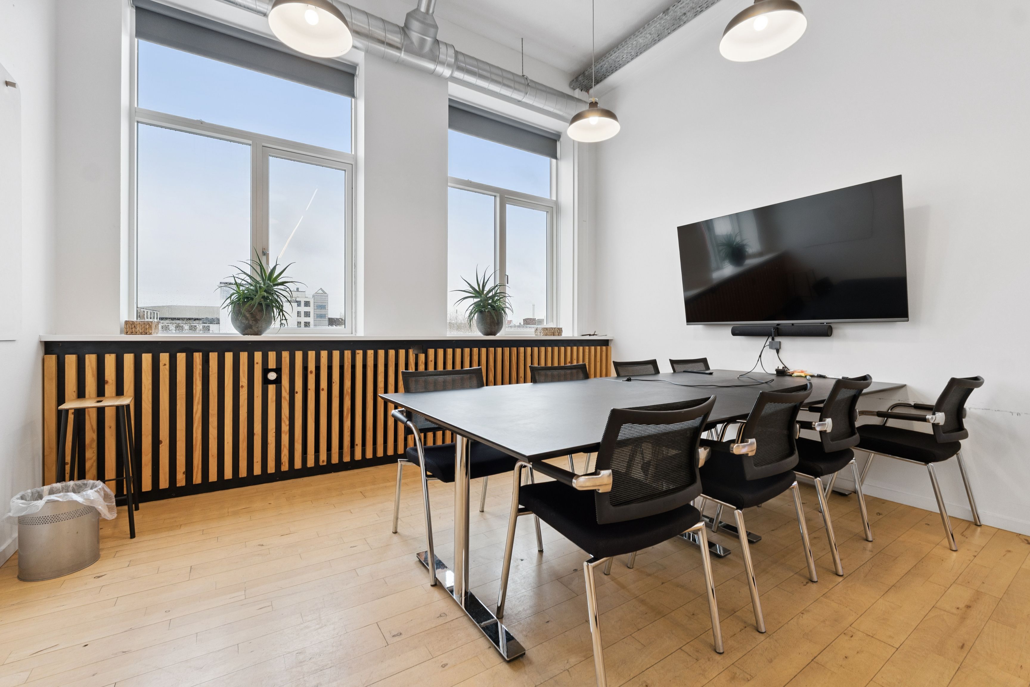 Kontorrum med lyse vinduer, moderne møbler og plantepoter på vindueskarm. Gulv med lyse brædder og stålbord med moderne stole. Loftslamper og et stort tv-apparat hængende på væggen