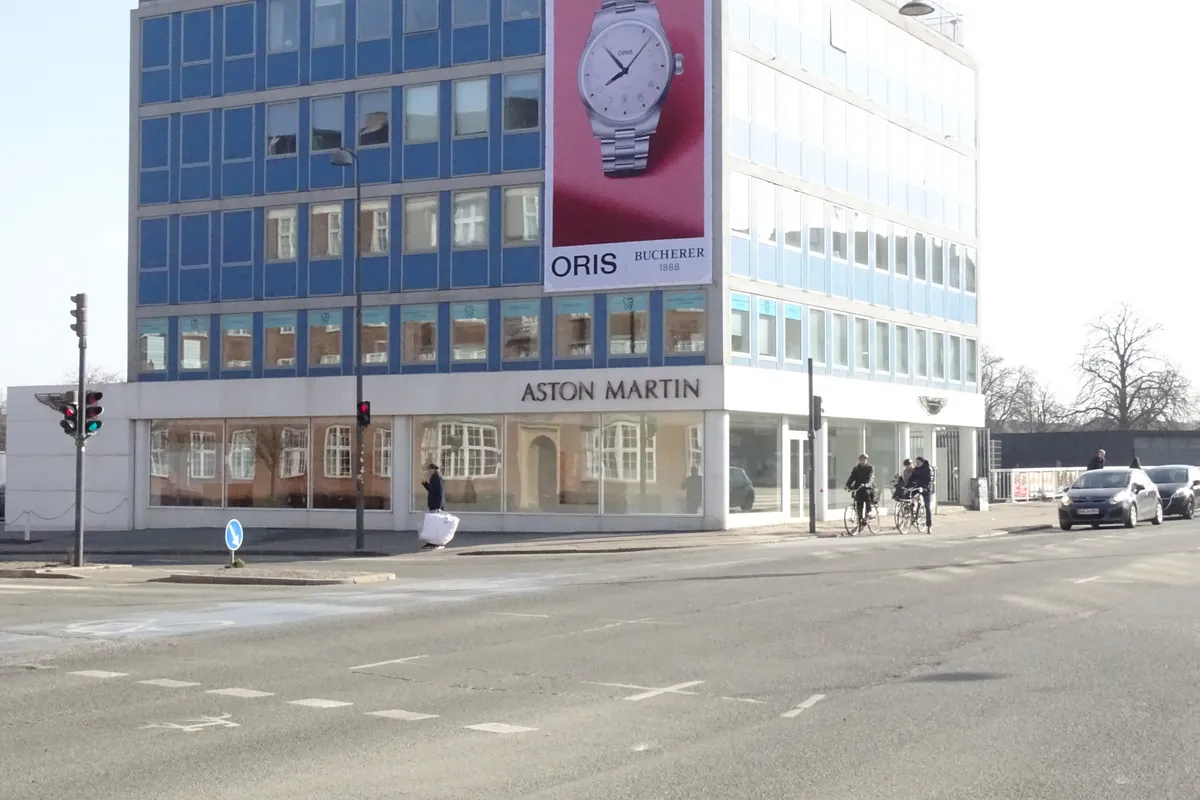 Mennesker kører med cykler og går forbi et luksusbutiksvindue med mærker som Aston Martin og Oris. Vej og trafiksignaler er synlige i baggrunden