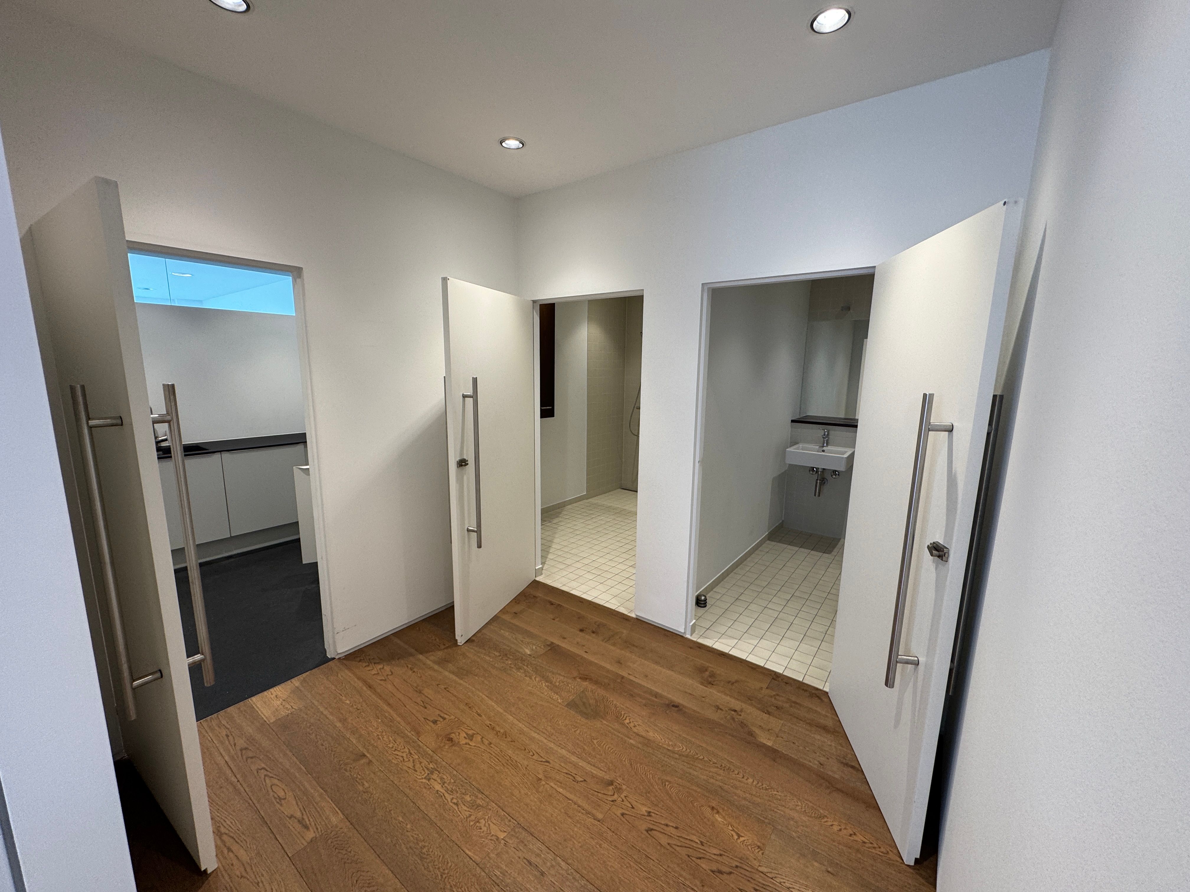 Et eksklusivt showroom med åbne hvide døre, lyse loftsbelægning og trægulve. Et luksusbadeværelse med hvide kakler og en hvid strygebrætte.