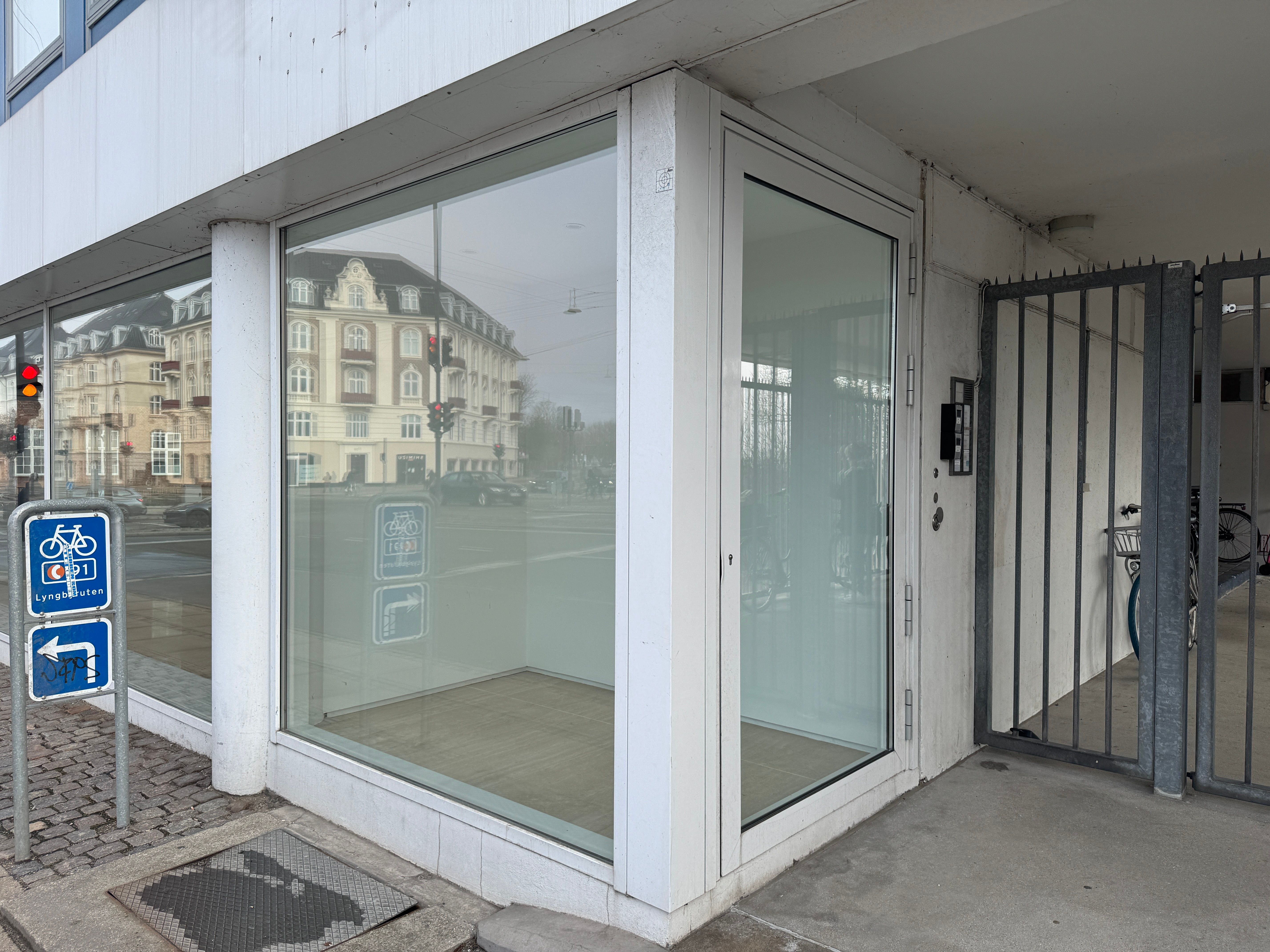 Strandvejen 6 i København Ø med en stor glasdør og et vindue, hvor der reflekteres bygninger og trafik. Der er parkeringsskilte for cykelparkering og trafiksignaler i baggrunden.