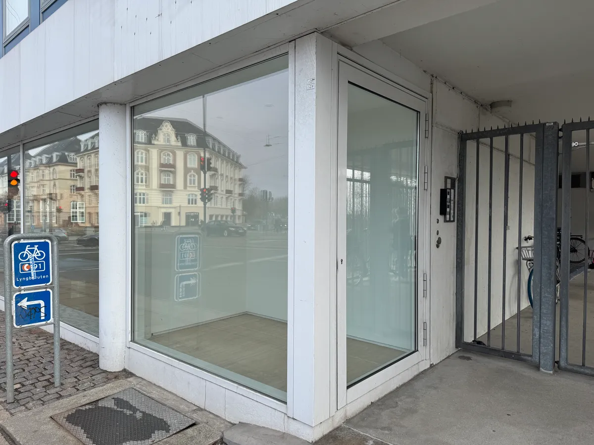 Strandvejen 6 i København Ø med en stor glasdør og et vindue, hvor der reflekteres bygninger og trafik. Der er parkeringsskilte for cykelparkering og trafiksignaler i baggrunden.