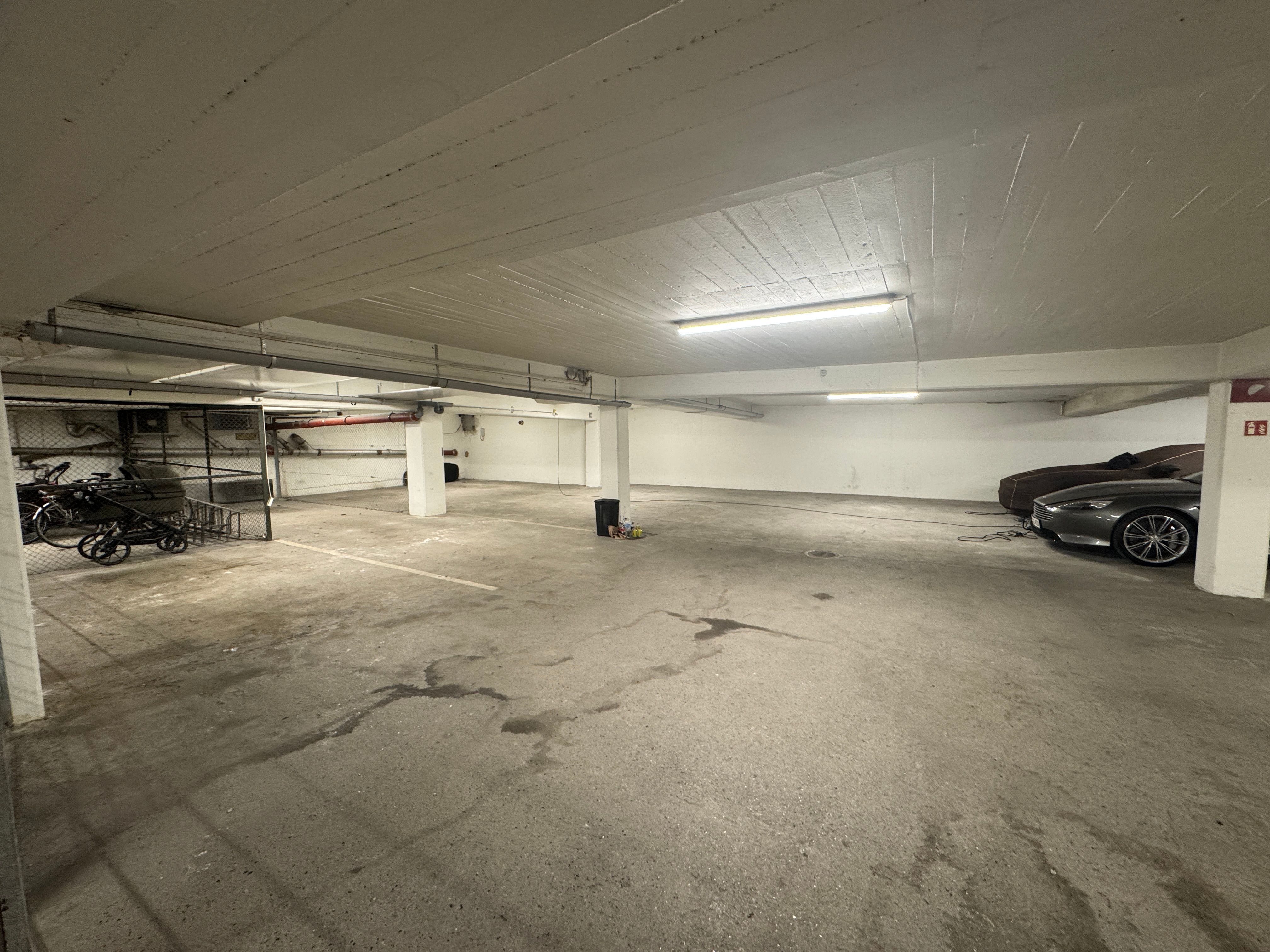 Garage med lyse højlofter og flere parkerede biler, en stående skraldespand og en cykel i en stor metalkurv