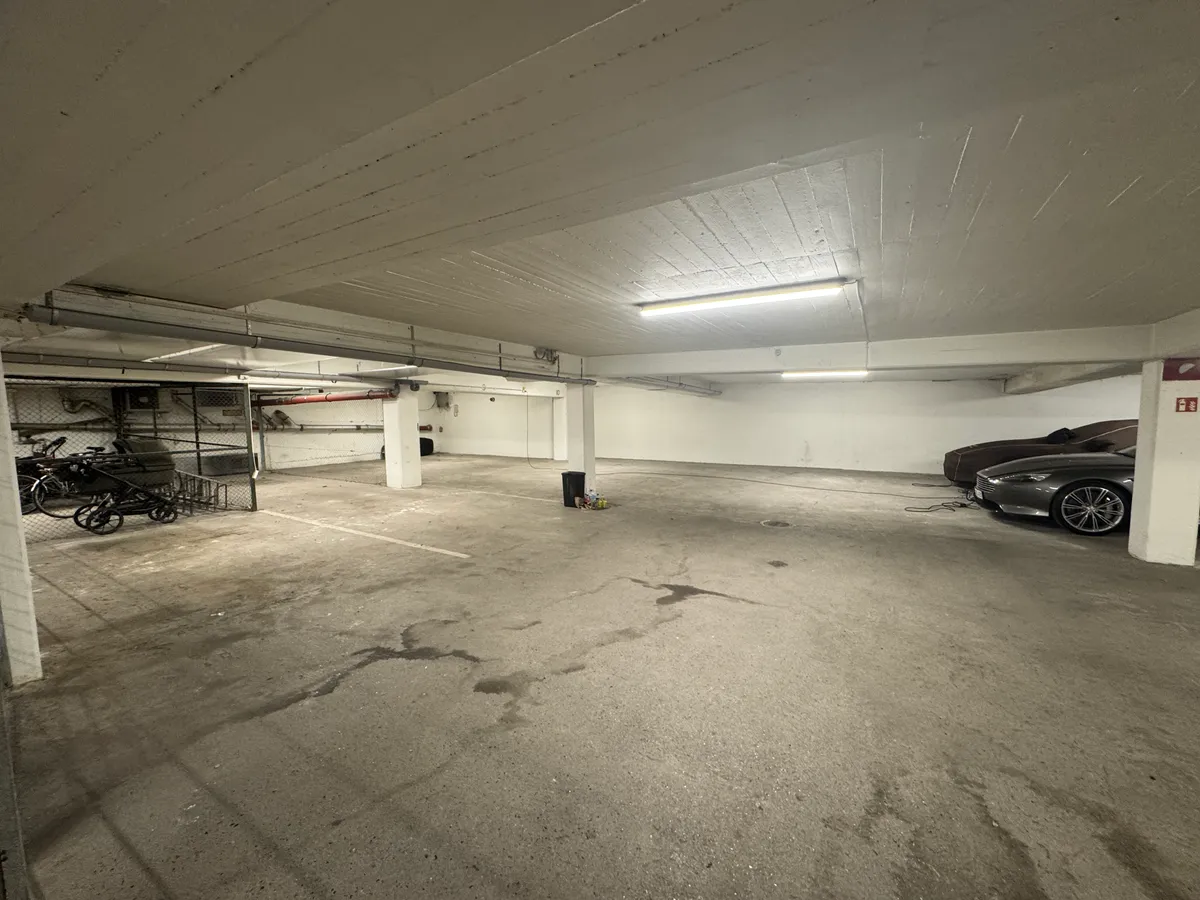 Garage med lyse højlofter og flere parkerede biler, en stående skraldespand og en cykel i en stor metalkurv