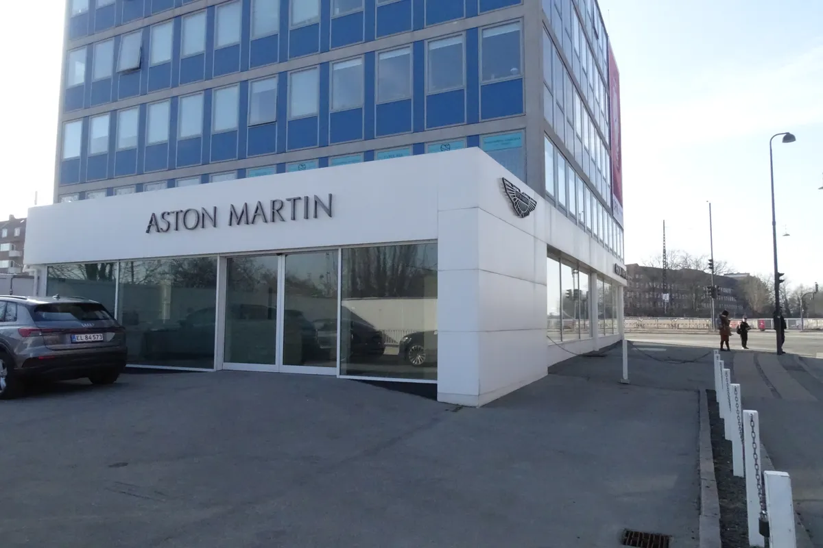 Forruden af en Aston Martin butik med logoet og firmanavnet Aston Martin. To personer går på fortovet og en person parkerer en bil.
