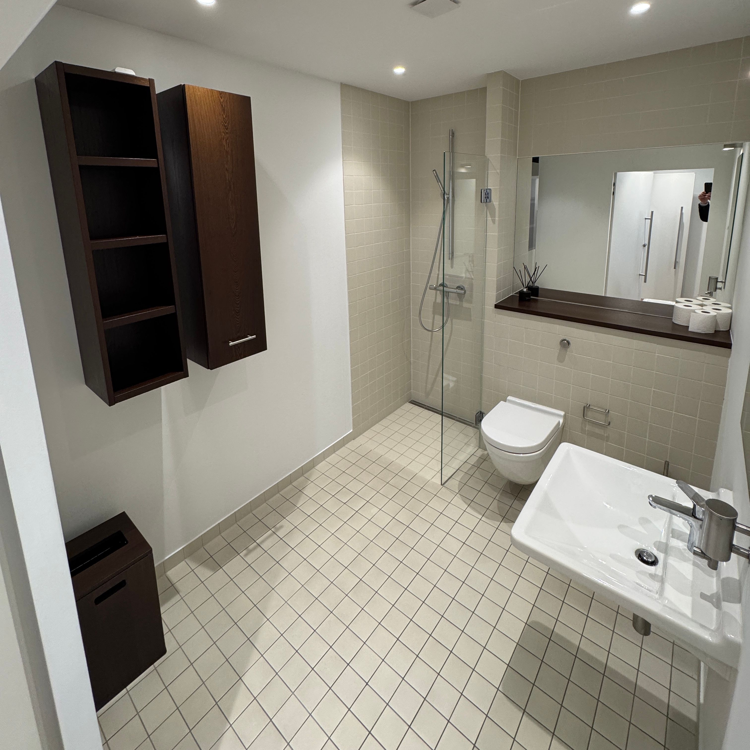 Moderne badeværelse med hvid flisegulv, luksus toilet, hængende toilethylde, og spejl med refleksion af et menneske.