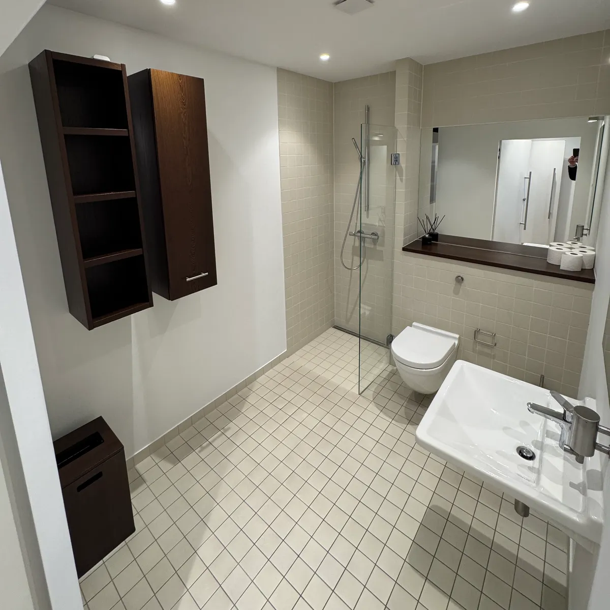 Moderne badeværelse med hvid flisegulv, luksus toilet, hængende toilethylde, og spejl med refleksion af et menneske.