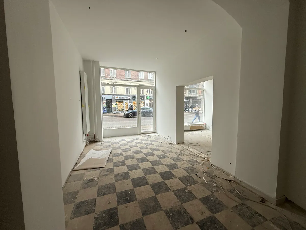 Udlejning af lokaler på Nørrebrogade 164 med udsigt til gågaden. En bil kører langs gaden og en person går forbi i udsigten. Lokalerne er i renovering.