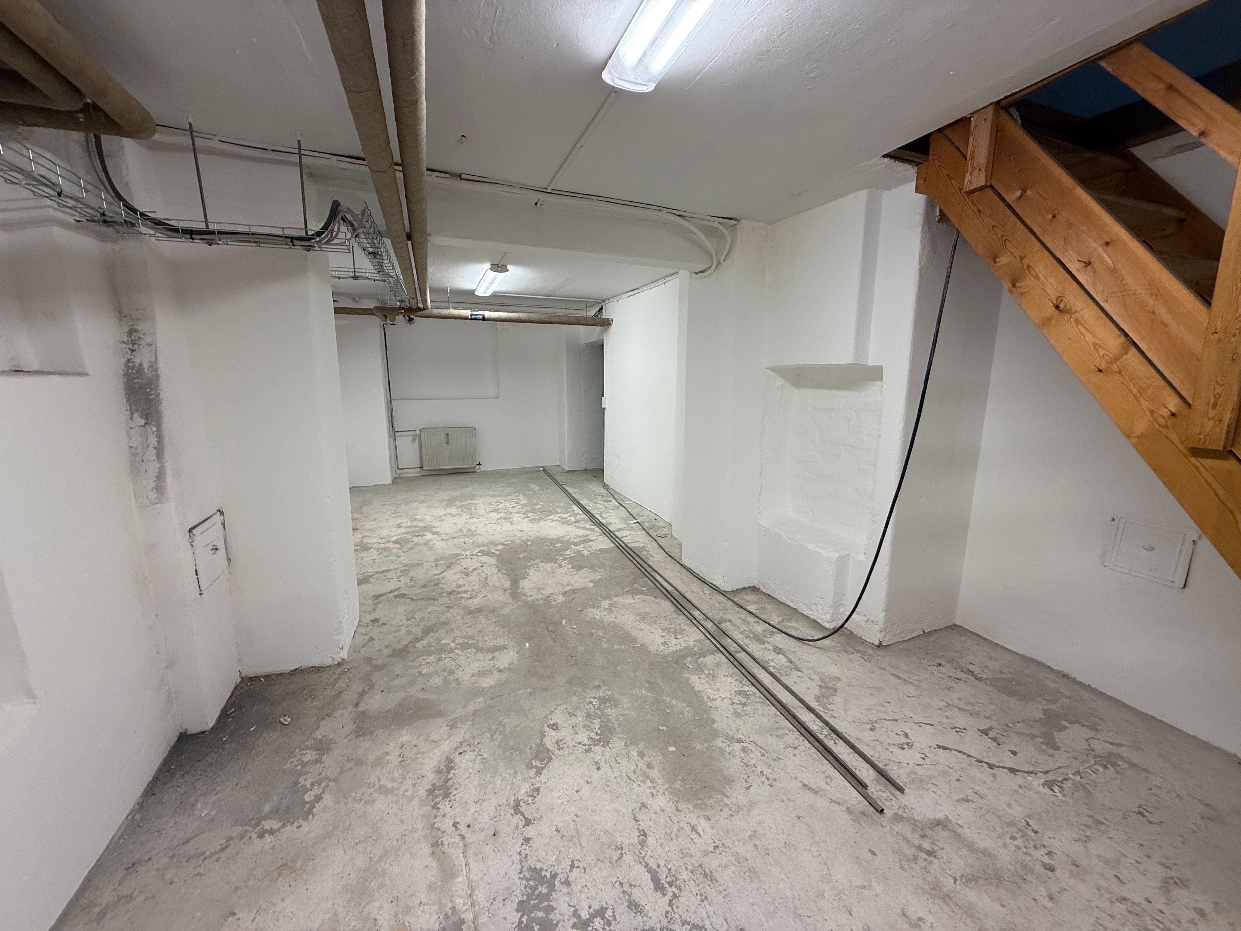 En tom og ubenyttet kælderlokale med lyse hvide vægge og hårdt cementgulv. To lange, svarte ledningsrør og en trætrappe i højre side.