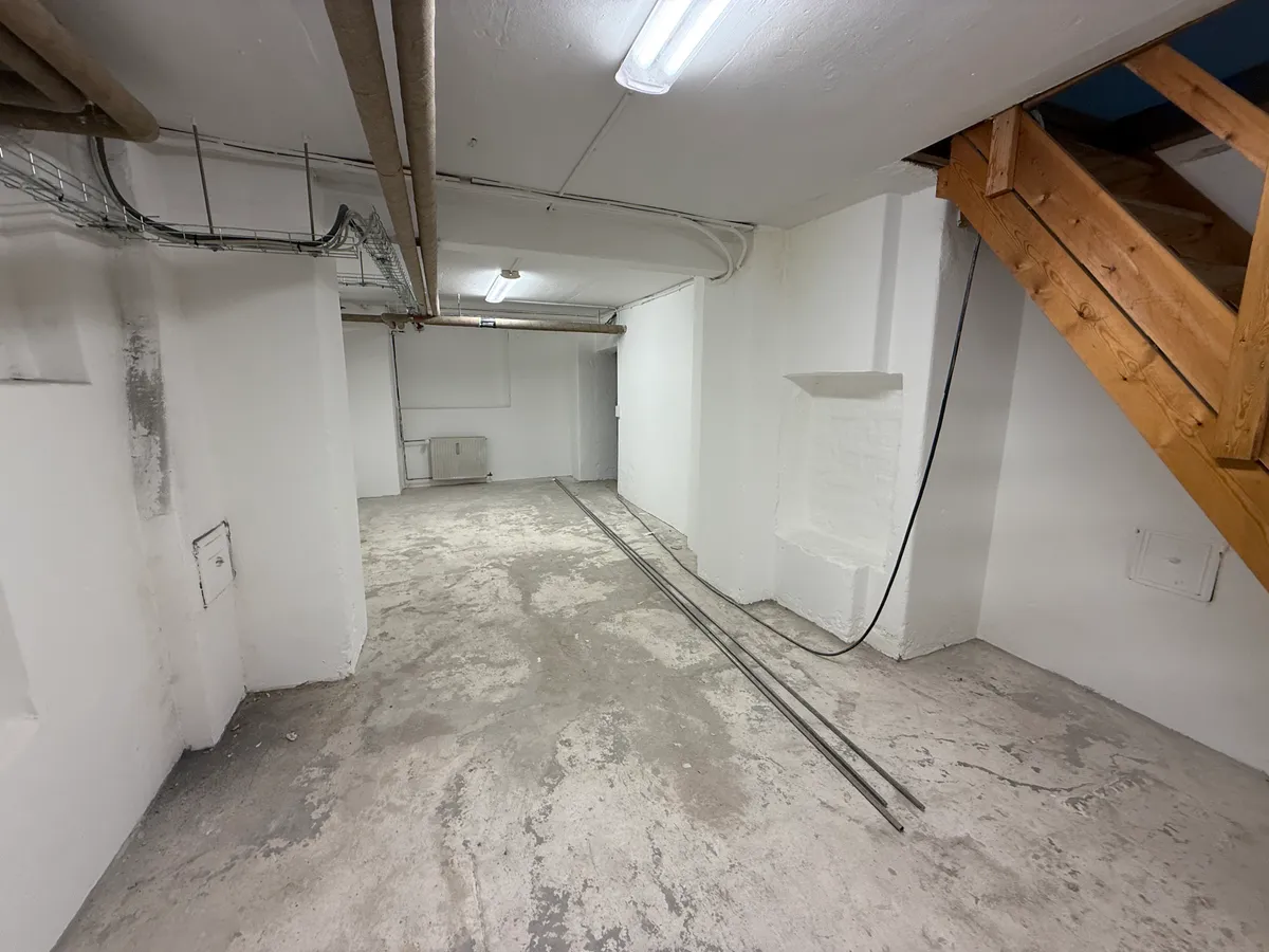 En tom og ubenyttet kælderlokale med lyse hvide vægge og hårdt cementgulv. To lange, svarte ledningsrør og en trætrappe i højre side.