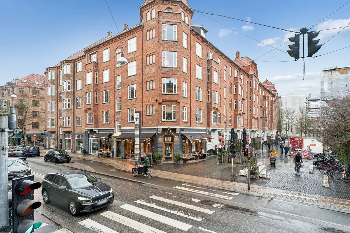 Stor bygning med røde mursten på hjørne. Personer går på fortovet og kører med cykel. Parkerede biler og trafiklys. Vejen er våd og tomme cykelstativer står på siden