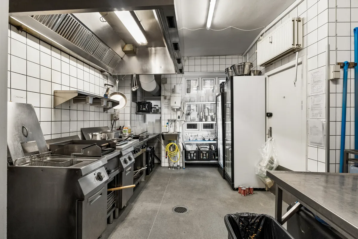 En lukket kommerciel køkken med køleskab, fryseskab og komfurer samt et skraldspand og en metalarbejdsbord. Væggene er hvide kakel og loftet er lyst.