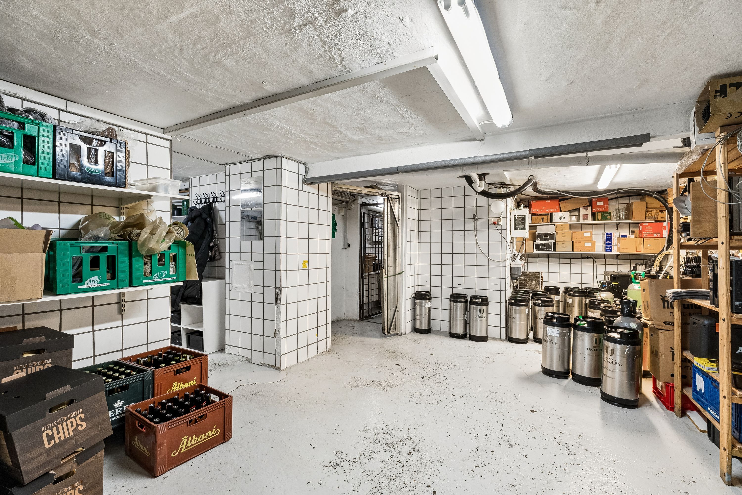 Lager med krydsfinerhyller, ølkrukker og køleskabe. Kasser med chips og øl samt diverse emballage. Lamper og røgrøg og røgrøg på loftet
