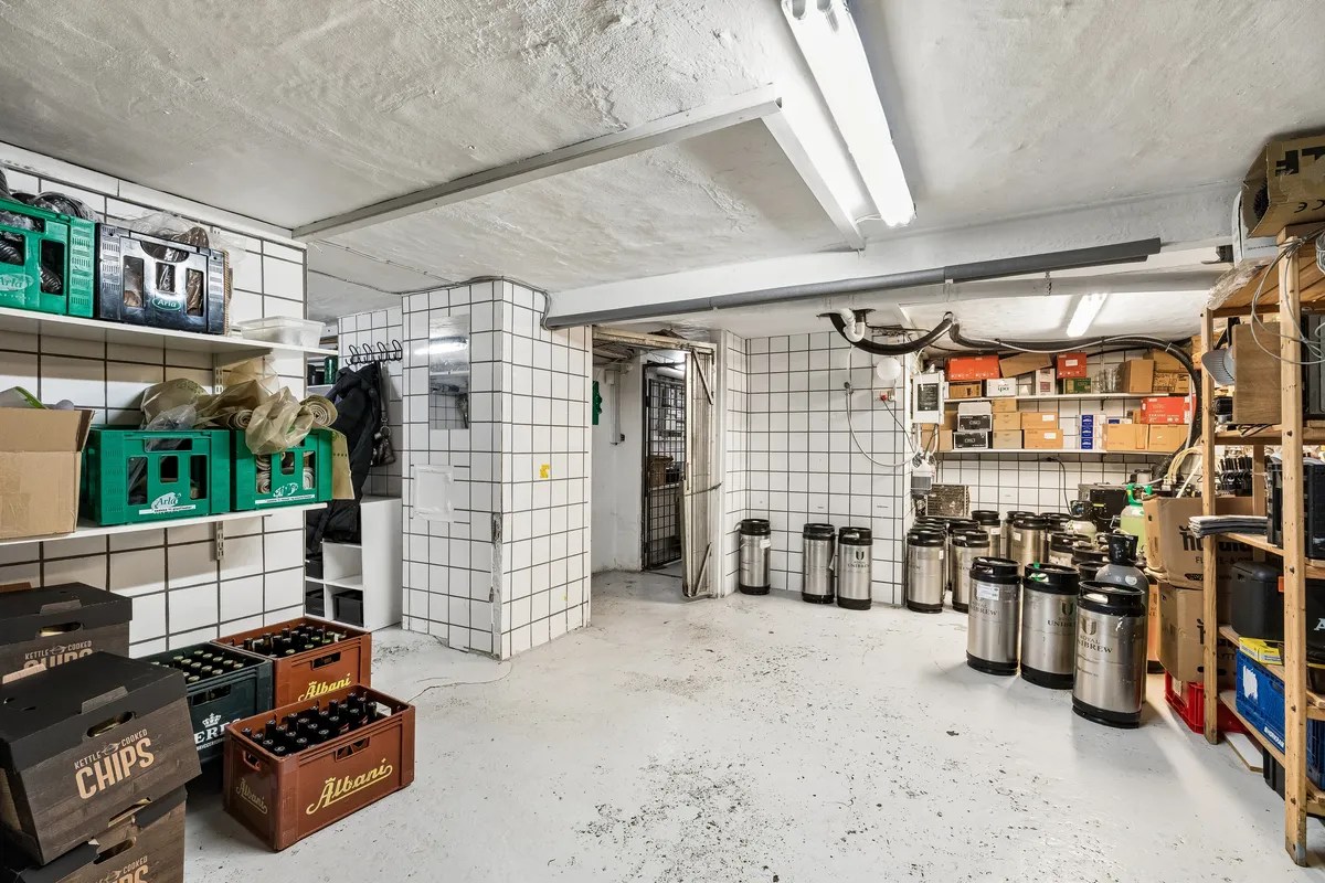 Lager med krydsfinerhyller, ølkrukker og køleskabe. Kasser med chips og øl samt diverse emballage. Lamper og røgrøg og røgrøg på loftet