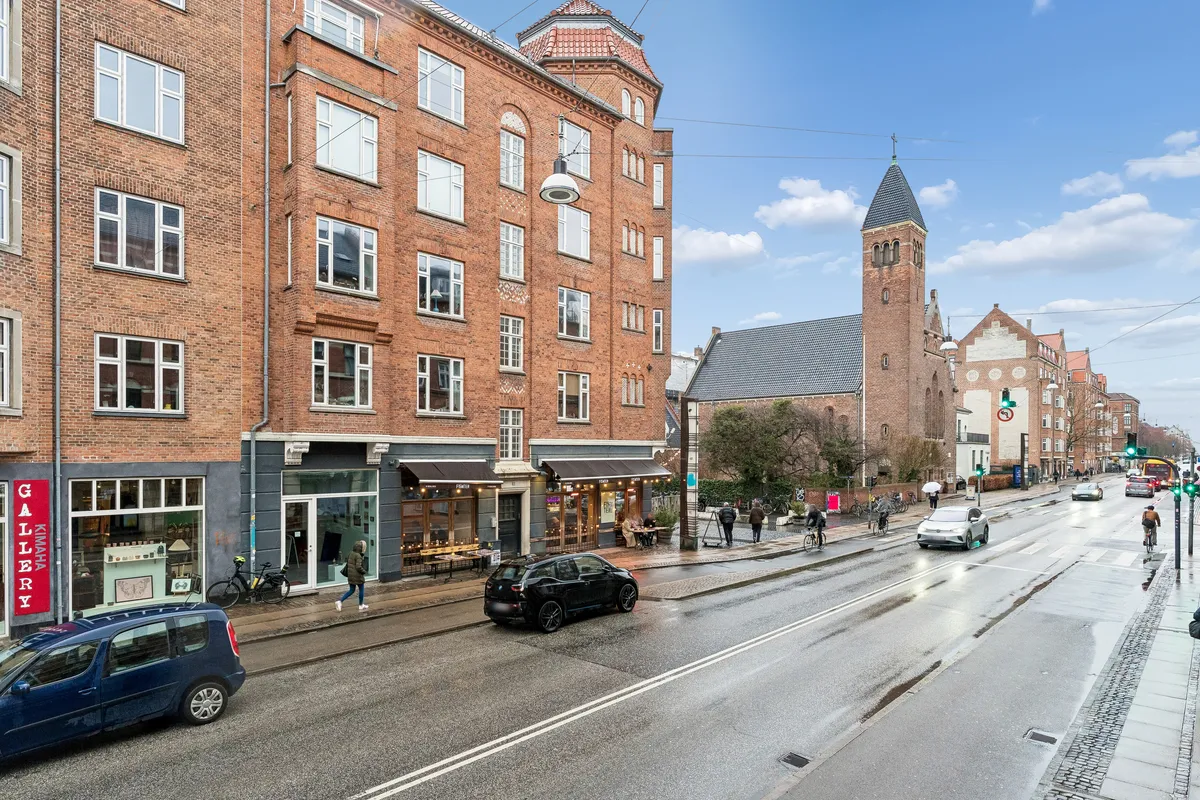 Butik og cafe i København S. En bil kører langs en gågade og krydser en vej, der er mærket med hvide streger. Mange mennesker går på fortovet foran en kirke