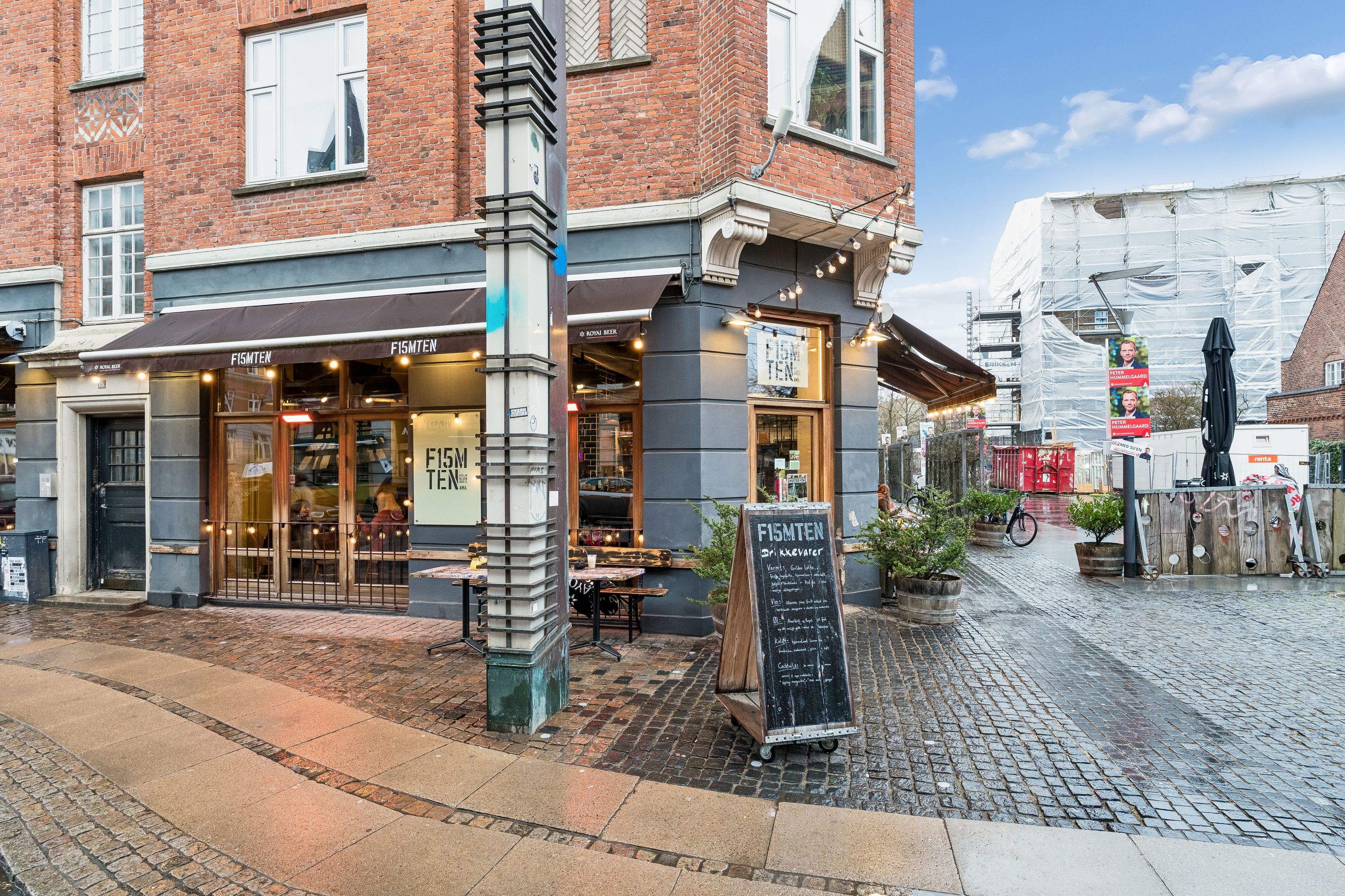 Restaurant Fiske Ten med skilte og solbeskyttelse udenfor, foran en bygning med røde mursten og vindue. En cykel og plantepotte står ved siden af. Der er stenbrolægget udenfor og bygningsudskæring og reklameplakater i baggrunden