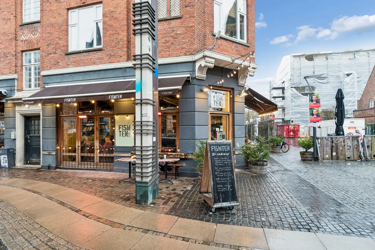 Restaurant Fiske Ten med skilte og solbeskyttelse udenfor, foran en bygning med røde mursten og vindue. En cykel og plantepotte står ved siden af. Der er stenbrolægget udenfor og bygningsudskæring og reklameplakater i baggrunden