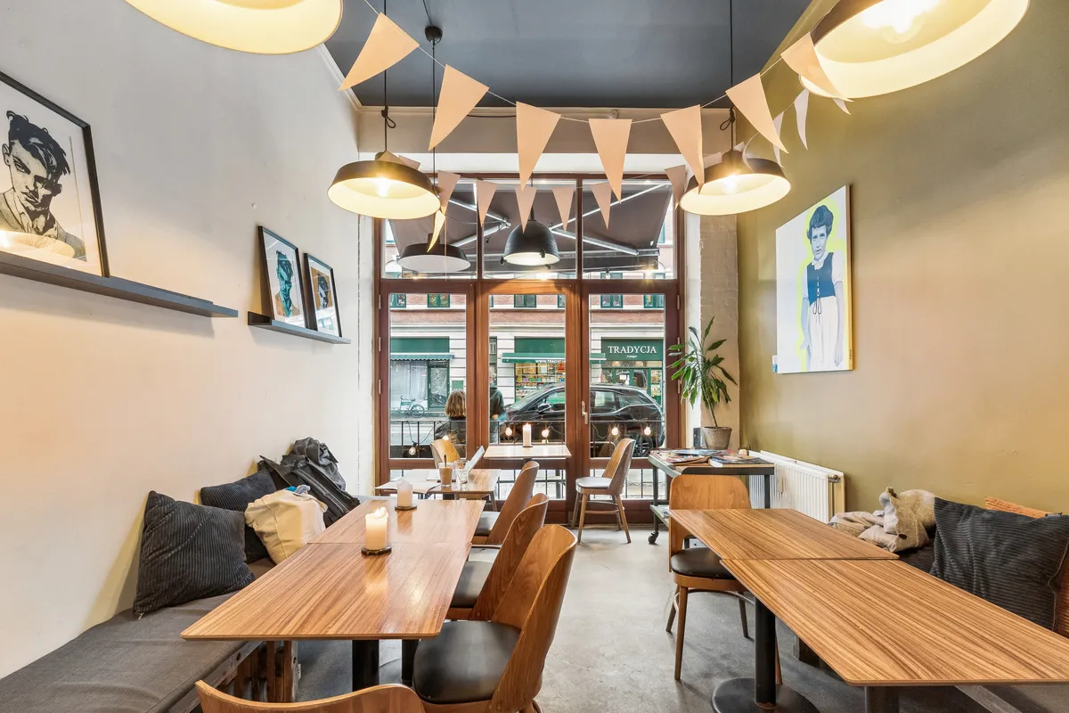 Restaurant med moderne interiør og puder på bænke. Træborde og stole med en plante og malerier på væggen. Lamper hængende fra loftet og vinduer med udsigt til biler og gågader.