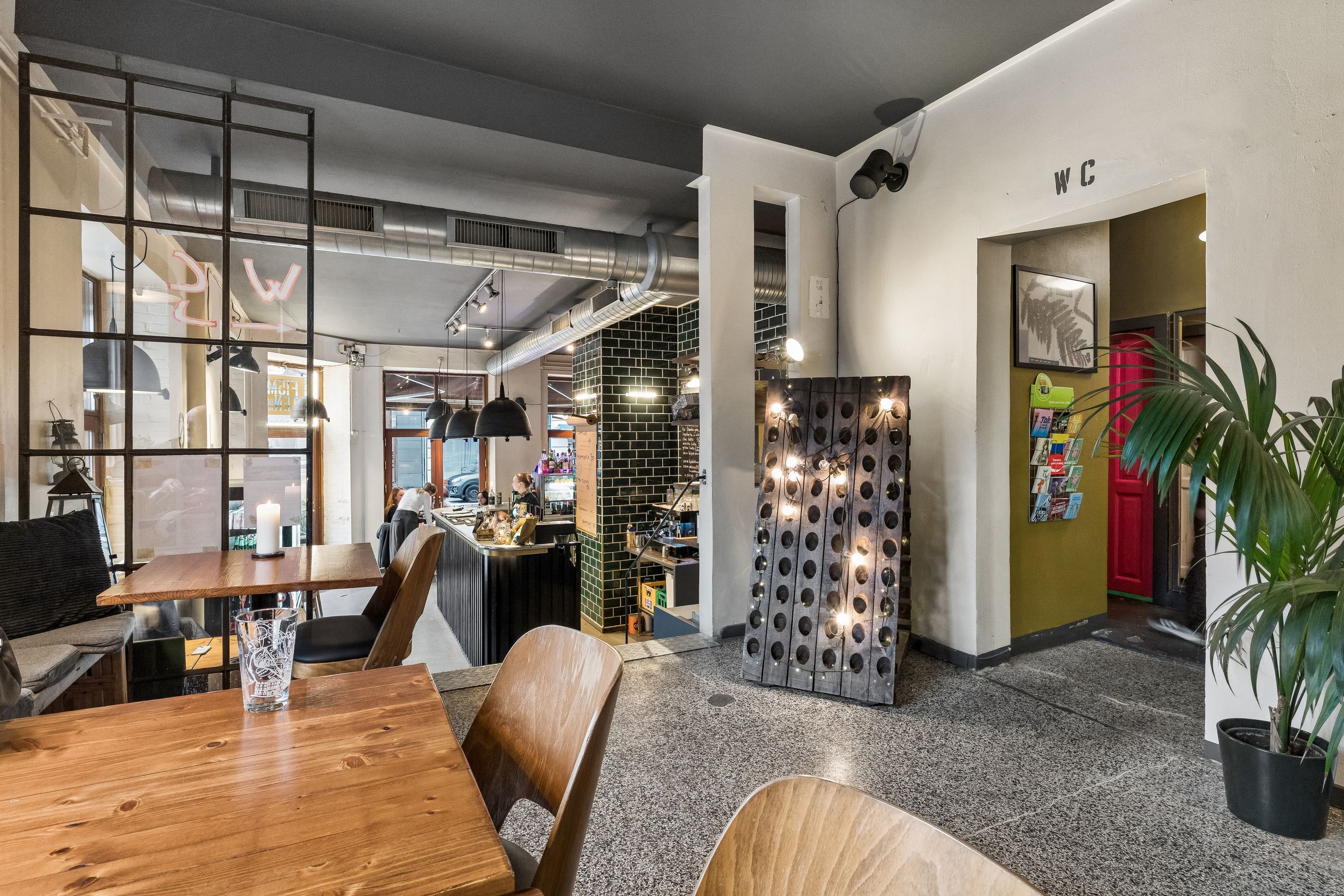 Inde i en restaurant med bord, stole og p-stand, der er dekoreret med lamper og plante. Væggene er dekoreret med billeder og de har en WC-skilt.