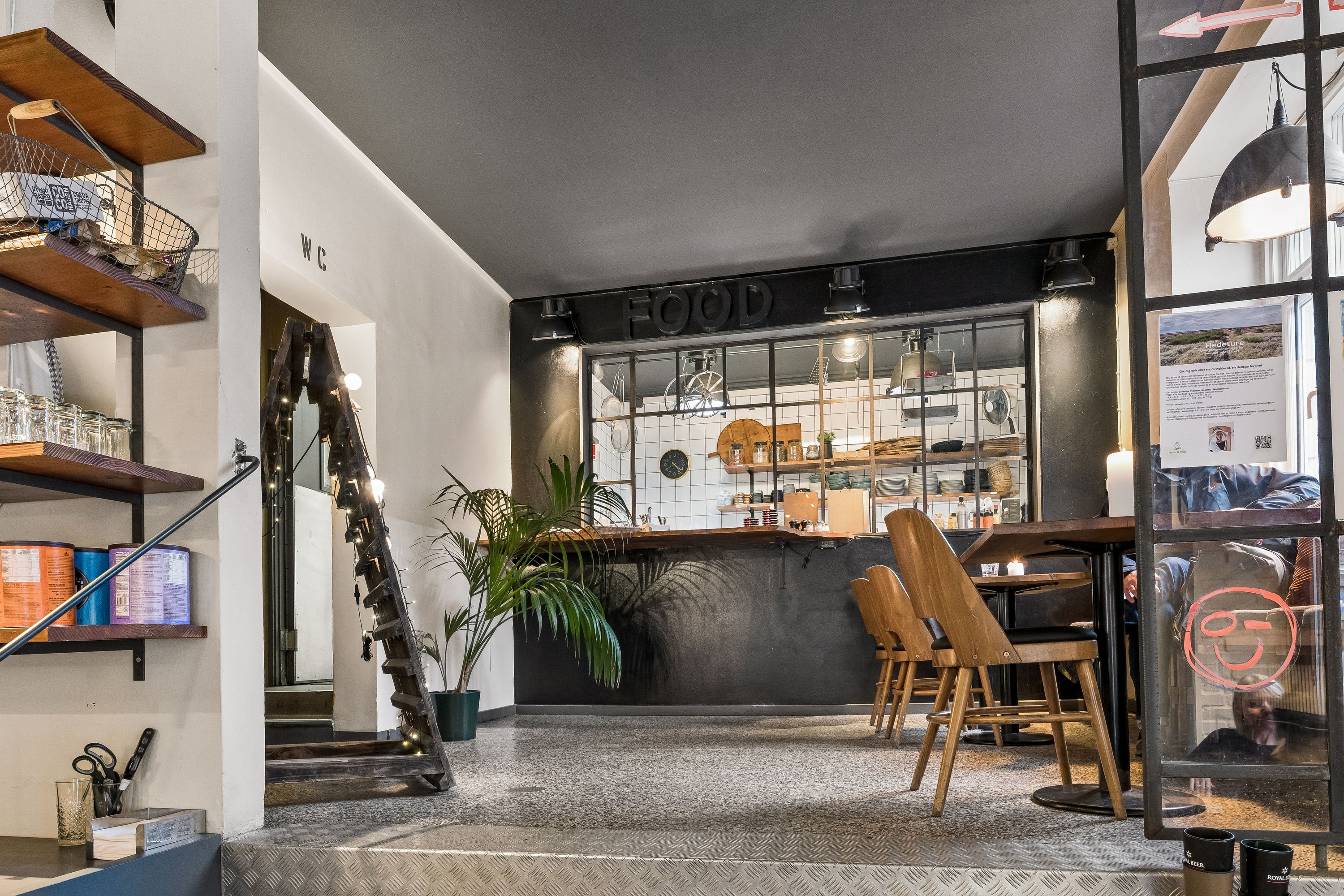 Forretningslokal i fuldt renoveret bygning med moderne indretning, lyse loftsvinduer og salgsområde med stolpebord og stole.