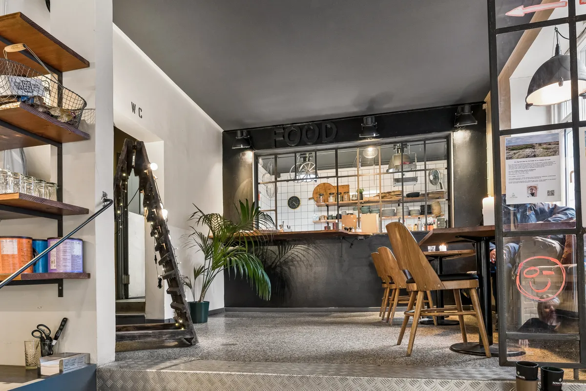 Forretningslokal i fuldt renoveret bygning med moderne indretning, lyse loftsvinduer og salgsområde med stolpebord og stole.