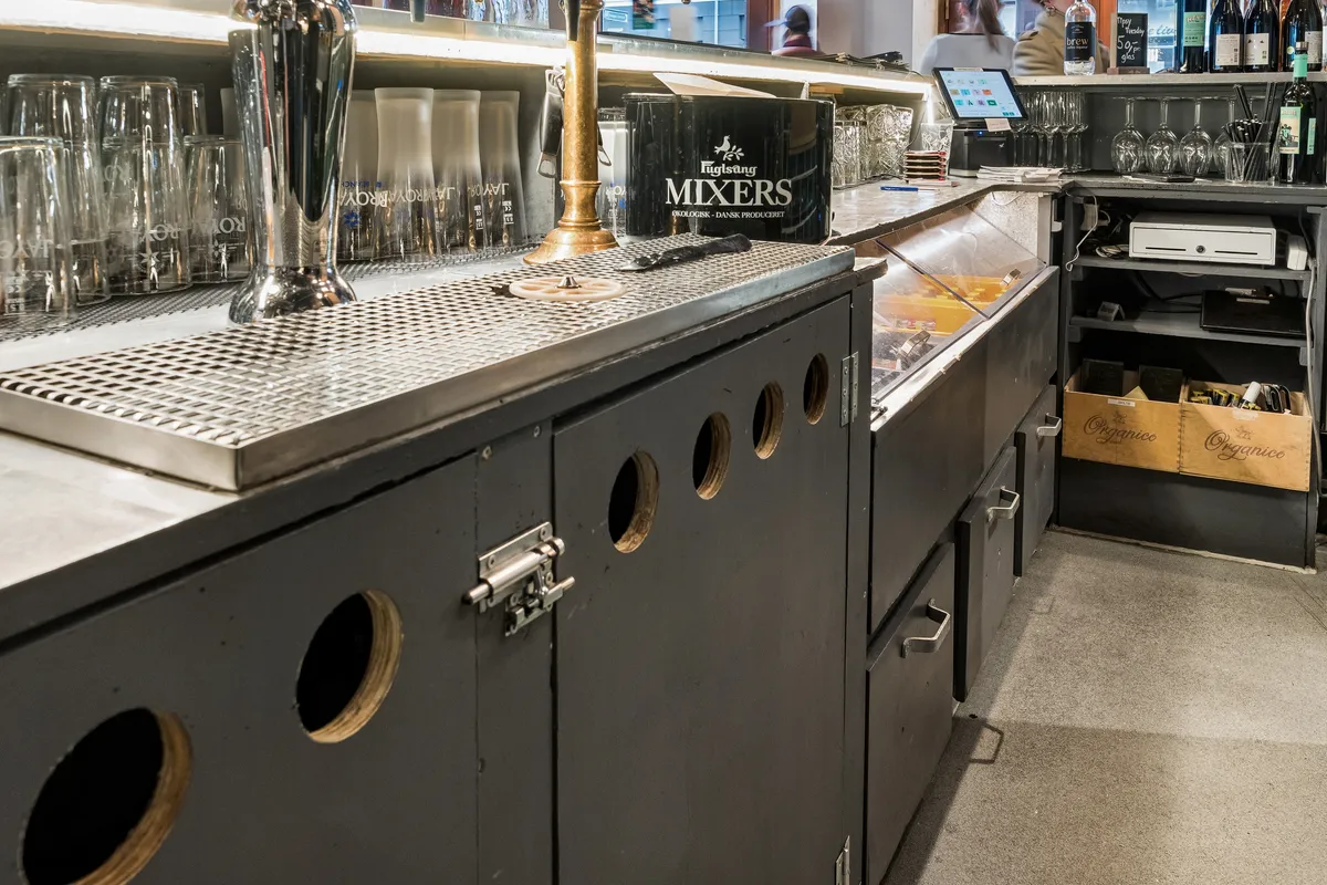 Restaurant med rustfri bar, glas, ølfat, mixers og vinflasker. Bar med skåle, tablet og øltønder samt køleskab med varer.