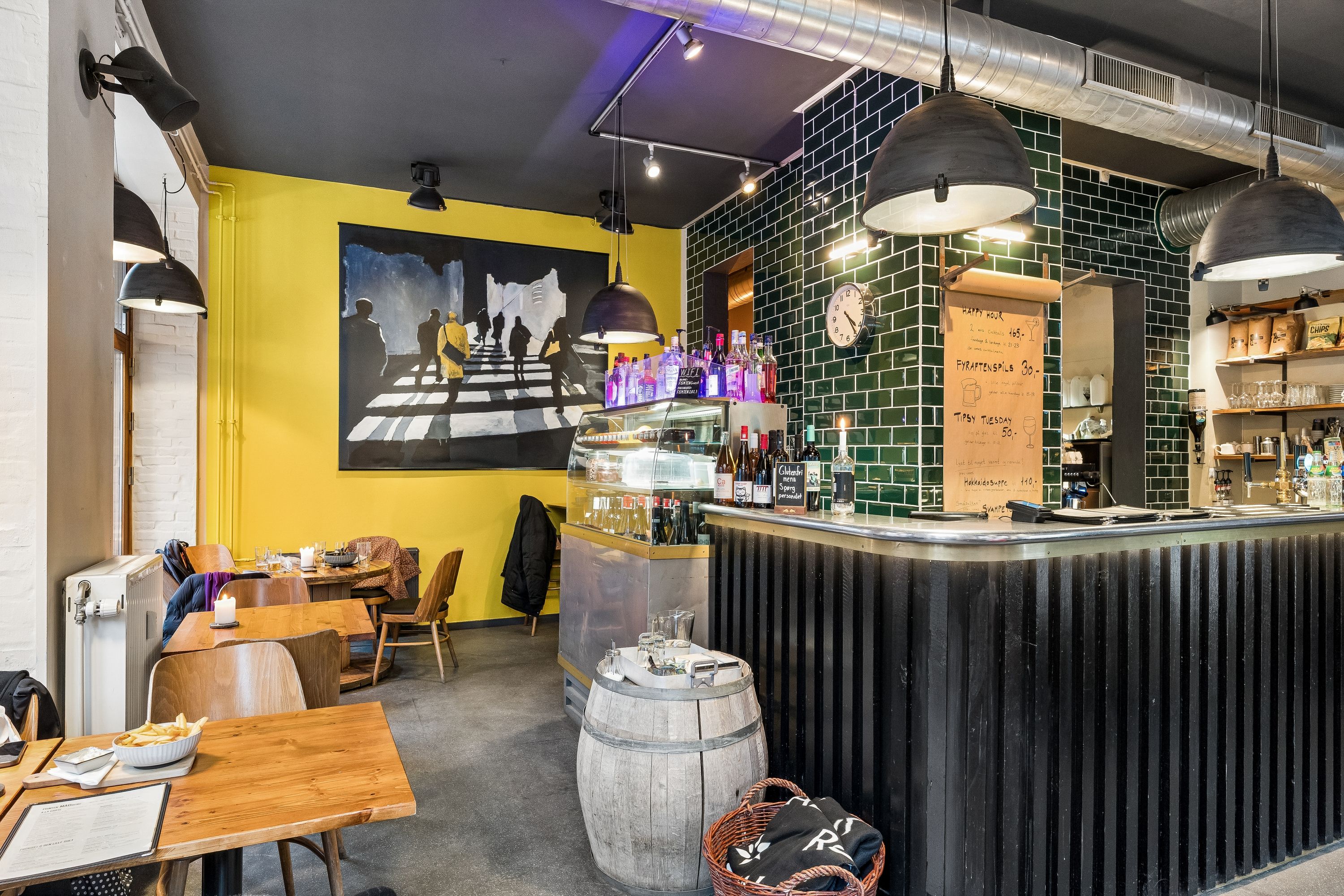 Restaurant med lyse gule vægge, træborde og stole, østeuropæisk maleri og flasker i udstillingsskabs. En stol med frak og papirhætte på gulvet og lysekroner i loftet