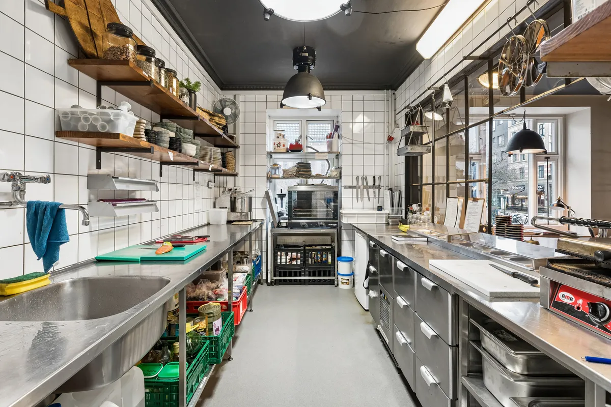 En professionel køkken med moderne indretning og udstyret med flere metalvogne og køleskabe. Der er en grydereol og flere ingredienser til madlavning på hylderne