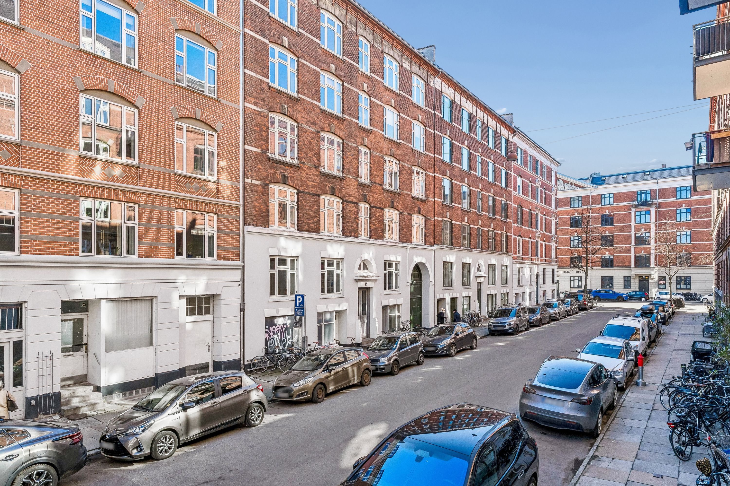 Marstalsgade 6, 2100 København Ø. Parkerede biler, bygning med teglstensfasade, to personer går ad gaden, cykler står parkeret, parkeringsskilt og grønne buske