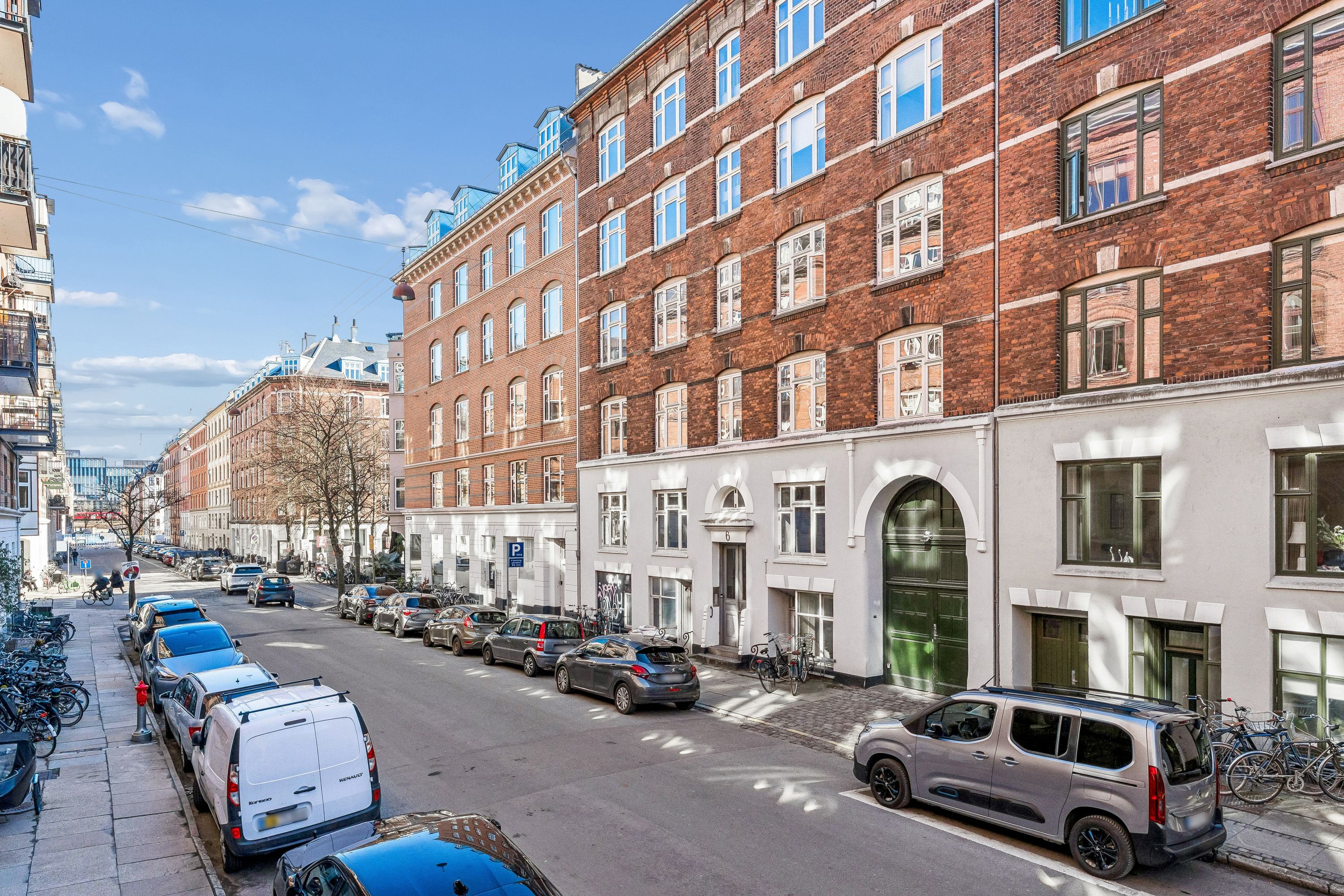 Flere biler parkeret på højre side af en bred gade med bygninger og mennesker, samt stel til cykler