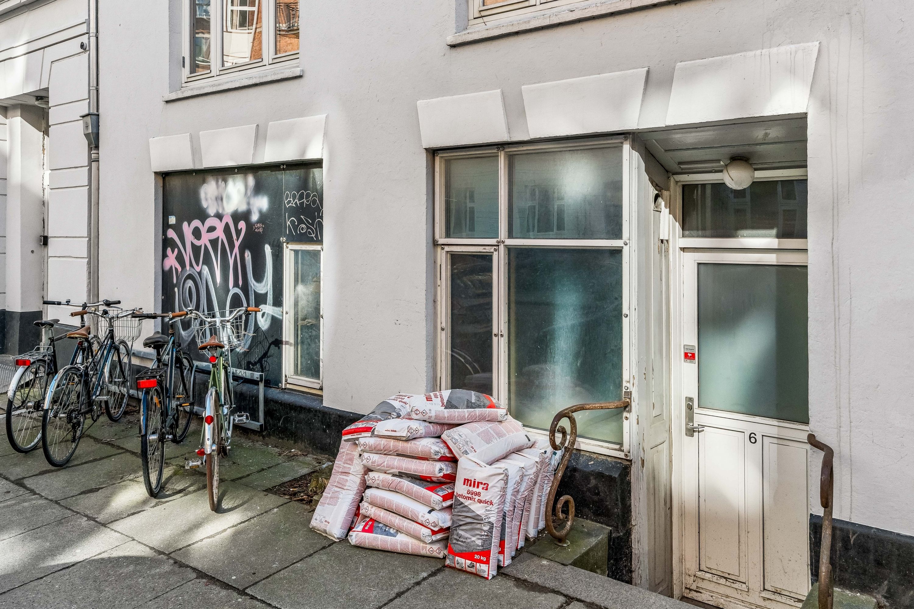 En hvid bygning med flere cykler parkeret og flere poser med Mira cement foran døren.