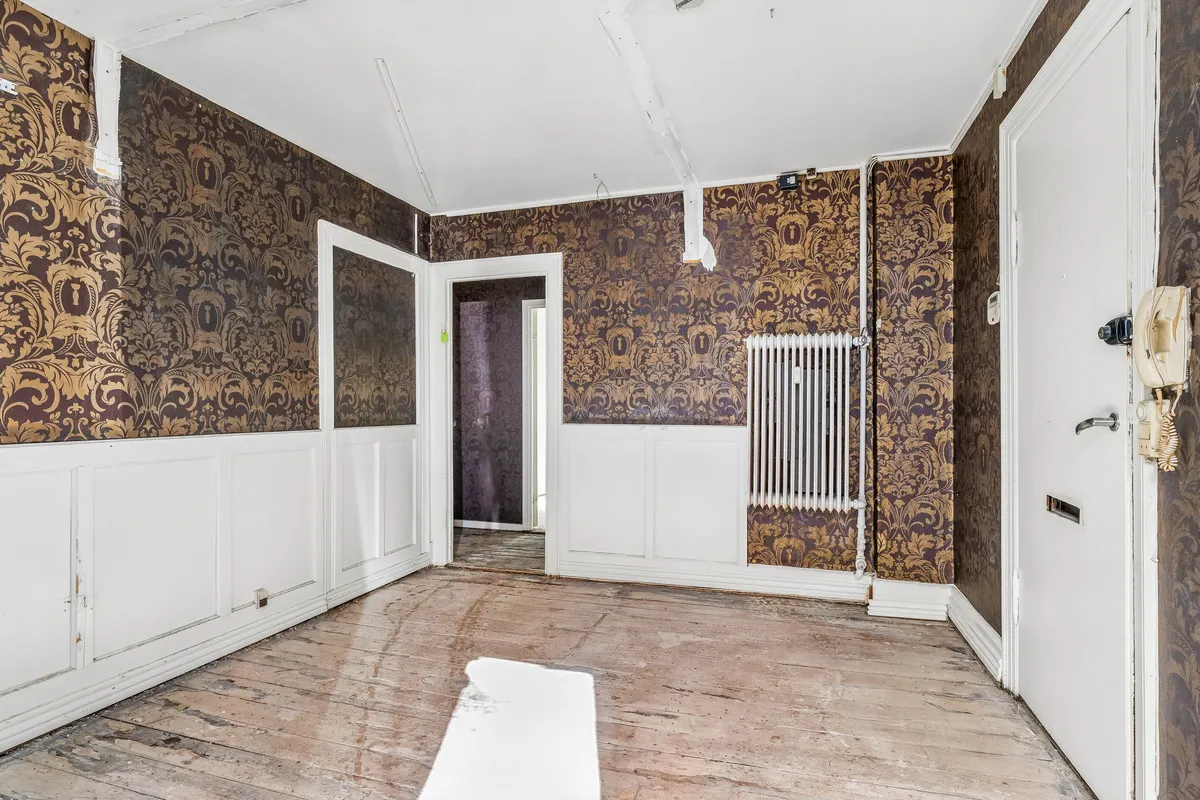En tom lejlighed med gamle mønstrede vægge, hvide paneler, lysegul gulv og gamle radiatorer. Døren er åben og der er en lys refleksion på gulvet
