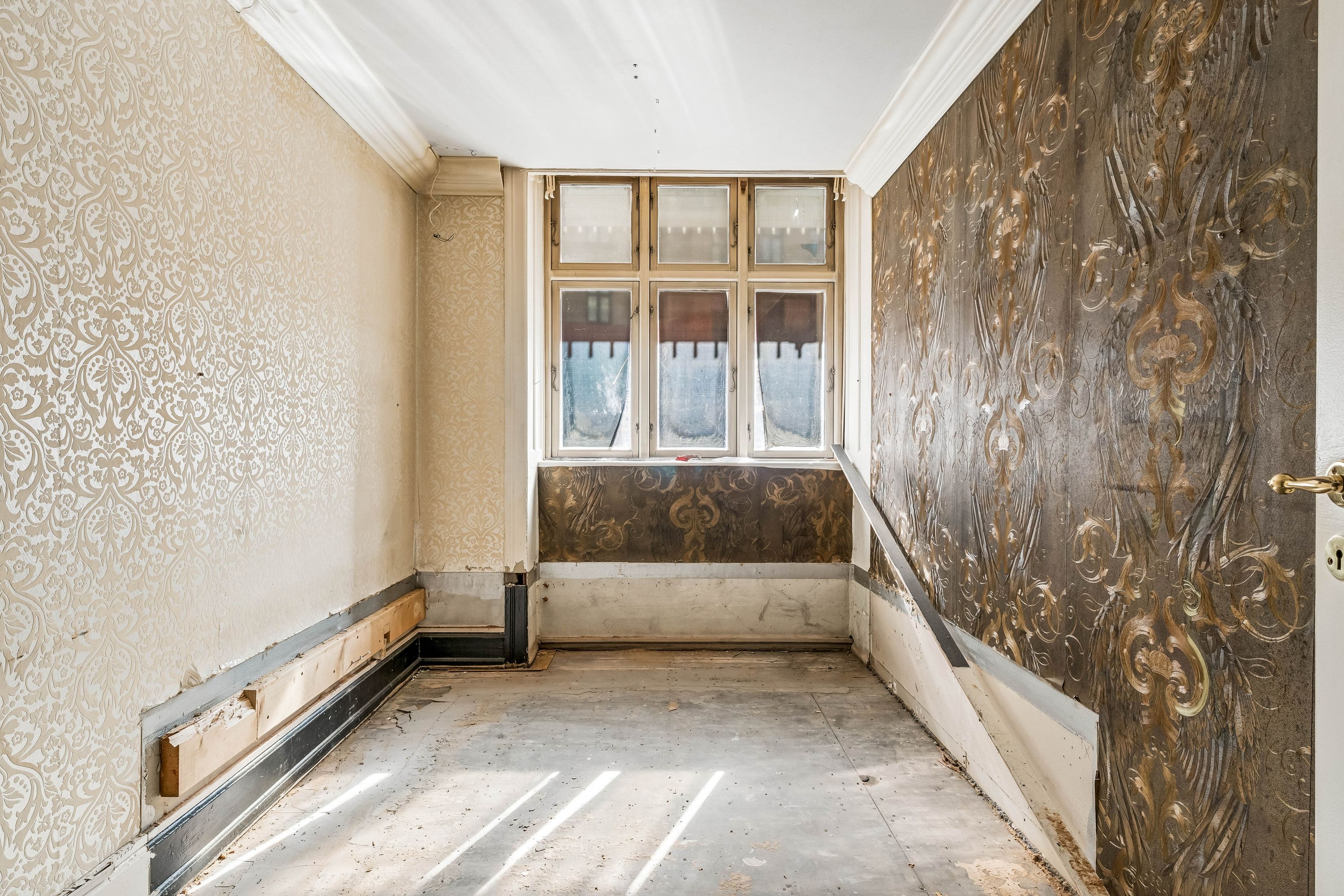 Tom og renoveringsbedyrvet husrum med lyse skikkelser og udvendige vægge i forskelige mønstre og materialer, og en vinduesbank med mønstret panel.