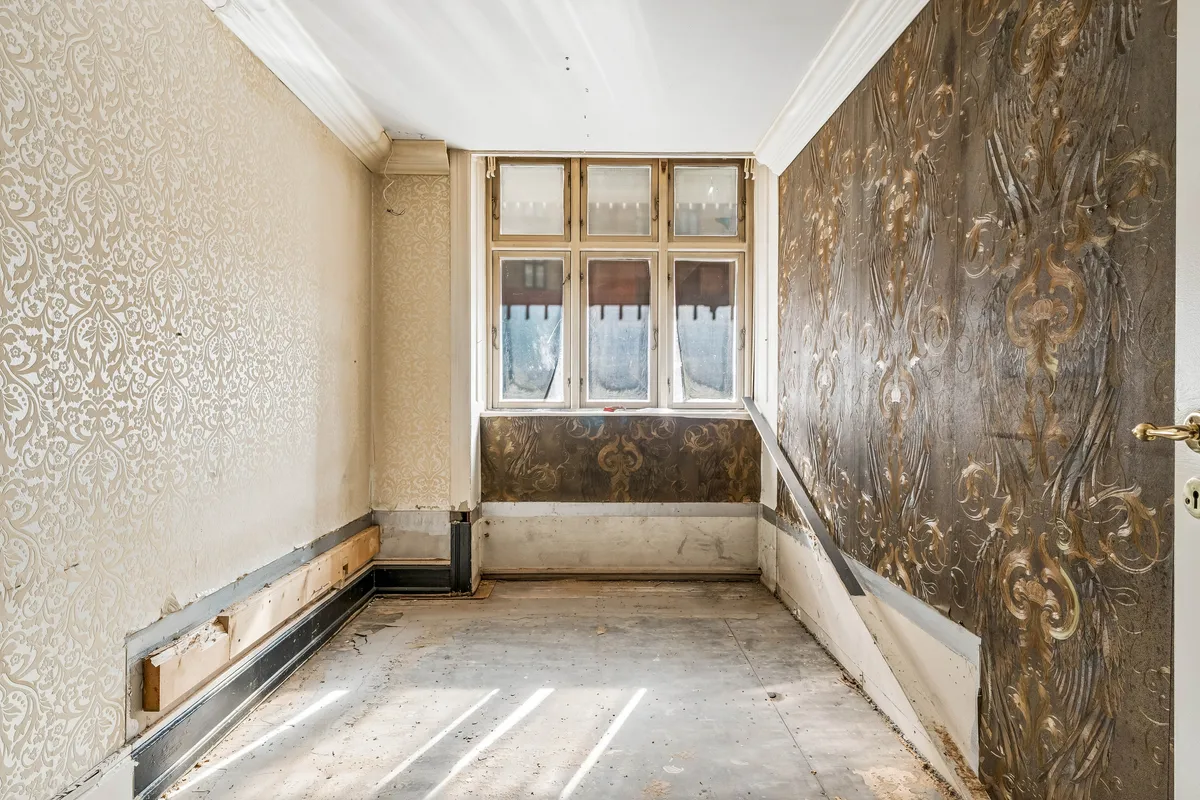 Tom og renoveringsbedyrvet husrum med lyse skikkelser og udvendige vægge i forskelige mønstre og materialer, og en vinduesbank med mønstret panel.