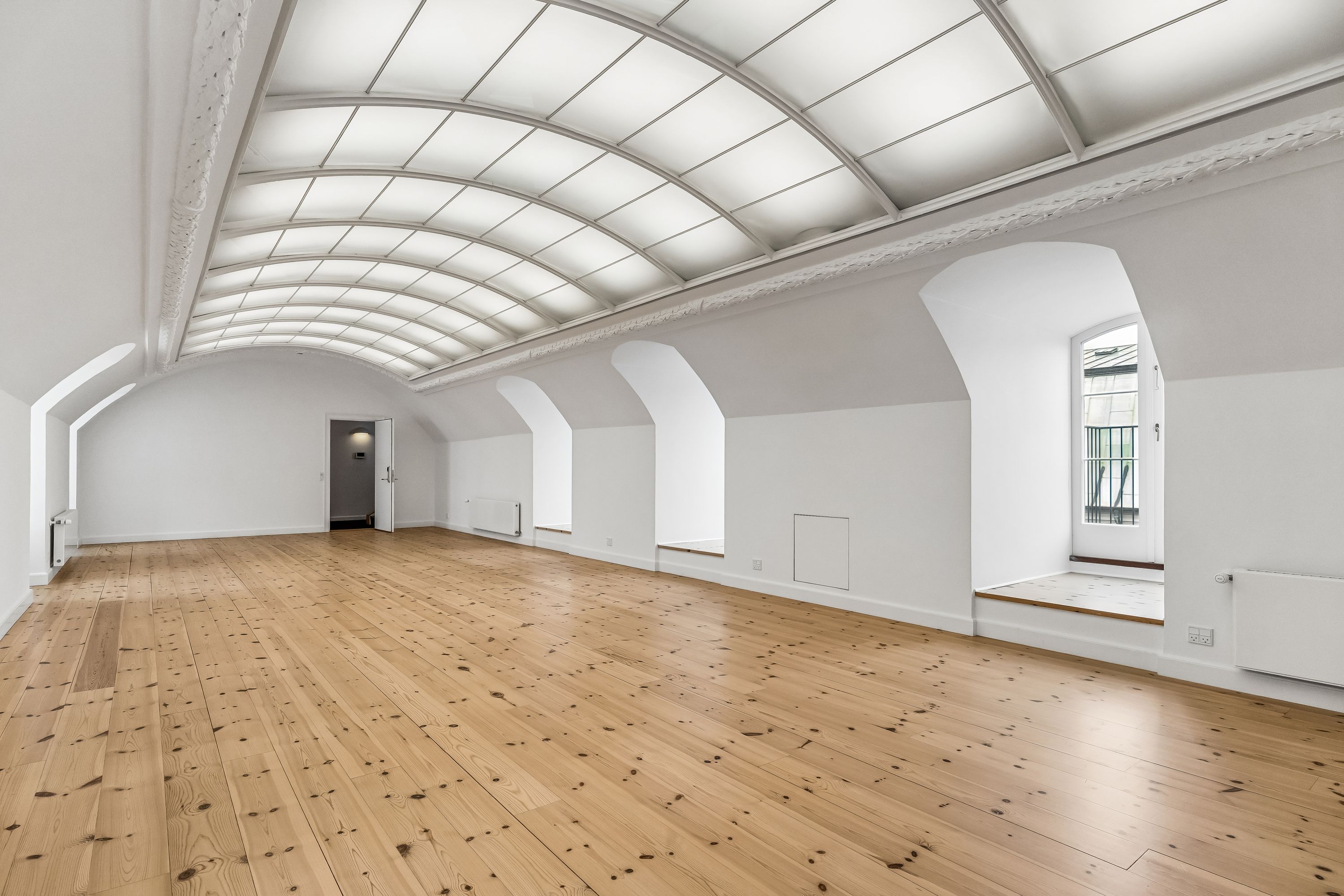 Kontor med højt hvide vægge og loft, lyse trægulve og vinduer med indbygget pergola. Værelse med åbne døre og lyseruder samt radiatorer