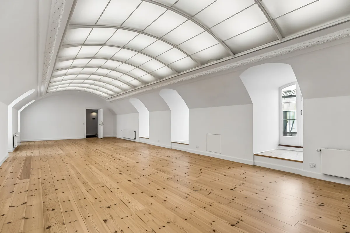 Kontor med højt hvide vægge og loft, lyse trægulve og vinduer med indbygget pergola. Værelse med åbne døre og lyseruder samt radiatorer