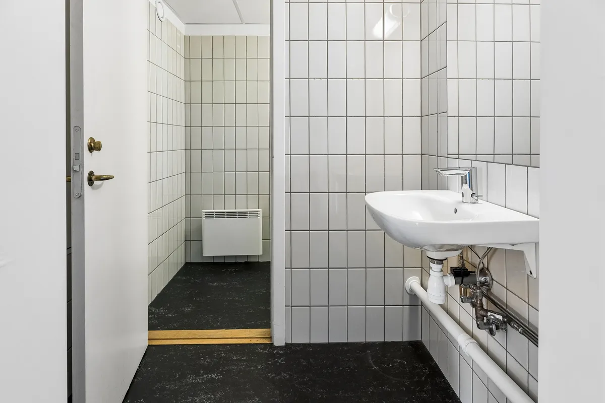 Rent kontor med hvidt fliserede vægge og gulv, hvor der er et rent hvidt vaskerum med både håndvask og skraldkurv samt et toilet med tørretumbler
