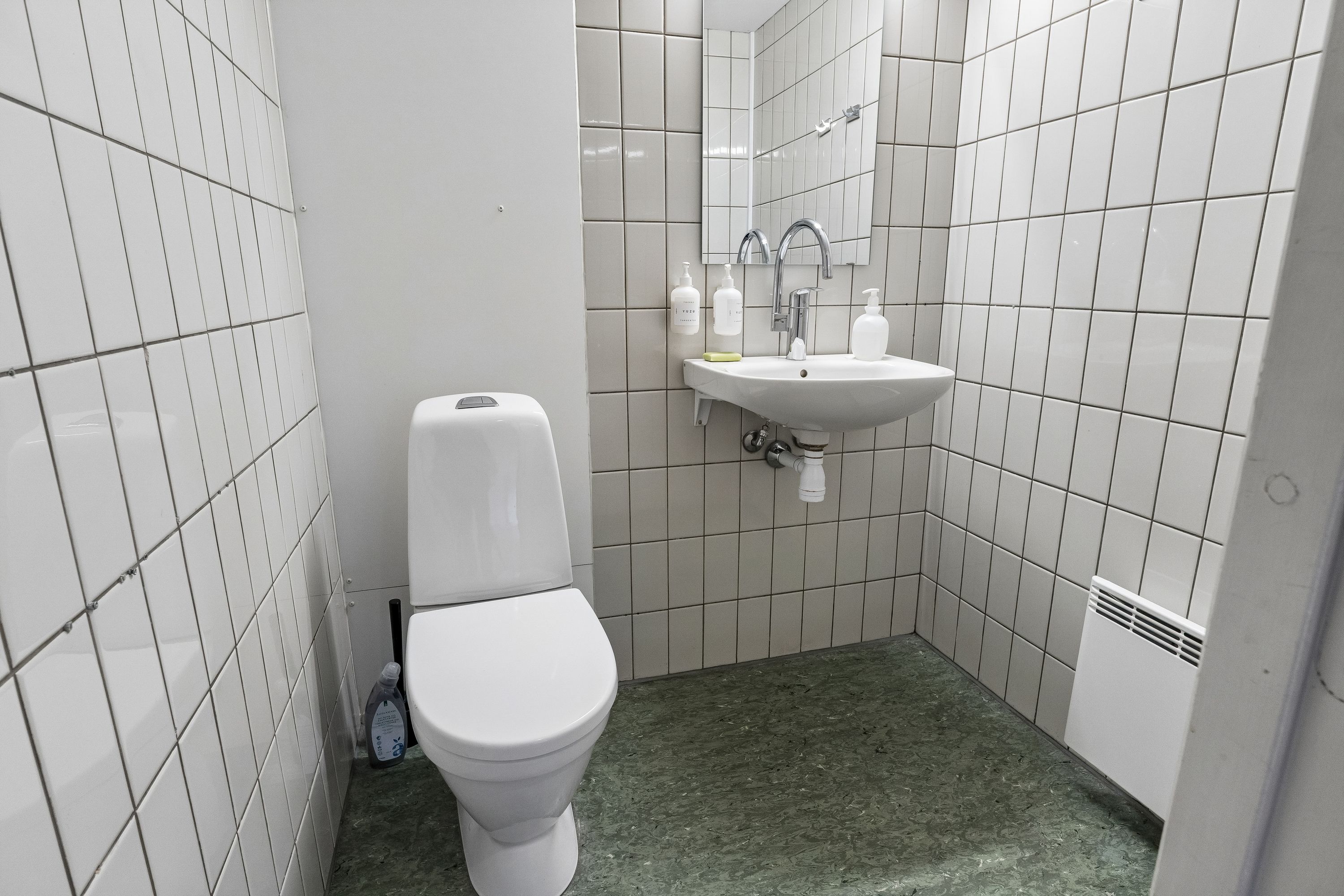 En lille badeværelse med hvide keramiske fliser og et toilet med toiletbørste, håndvask og sæbe i hvid skål samt et håndholdt håndklæde