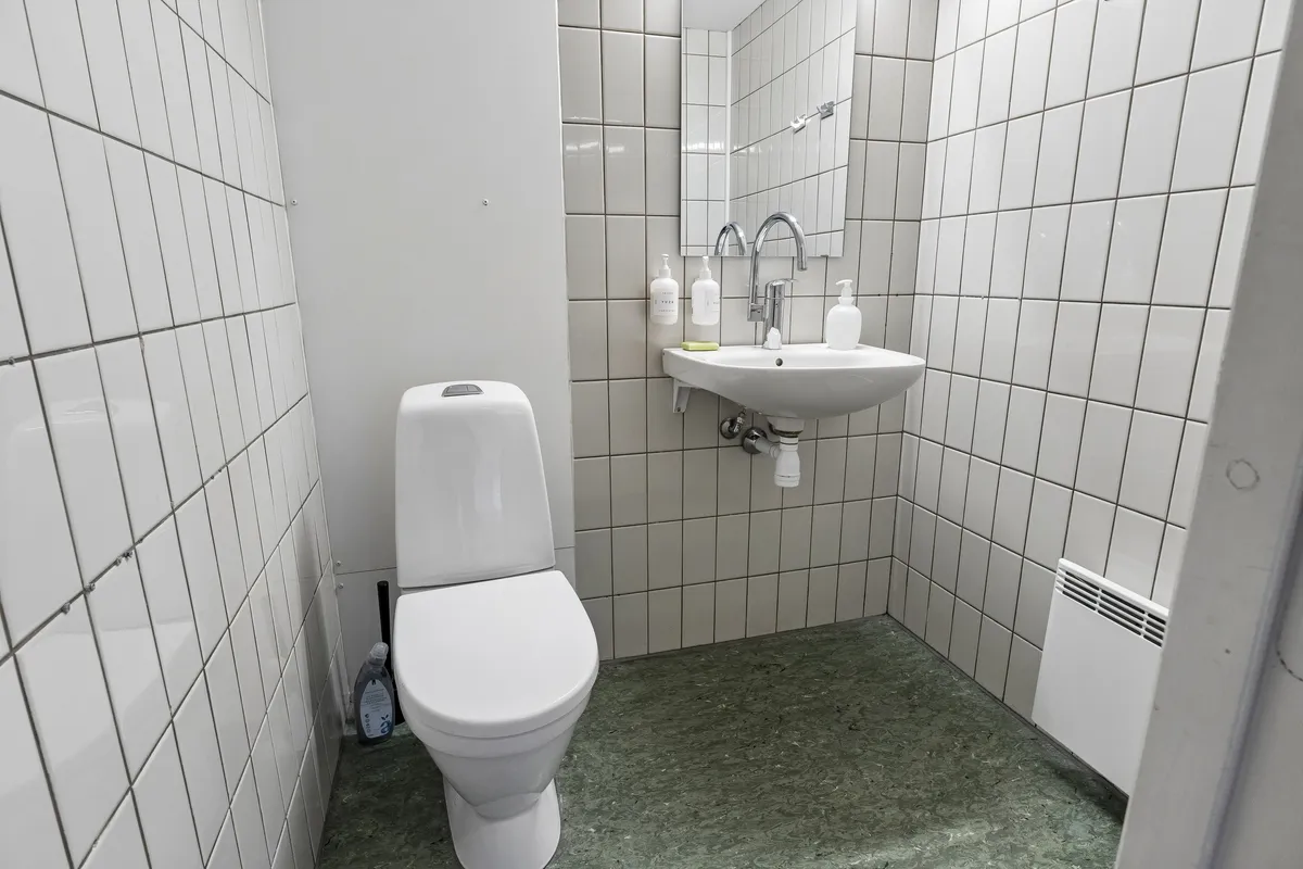 En lille badeværelse med hvide keramiske fliser og et toilet med toiletbørste, håndvask og sæbe i hvid skål samt et håndholdt håndklæde