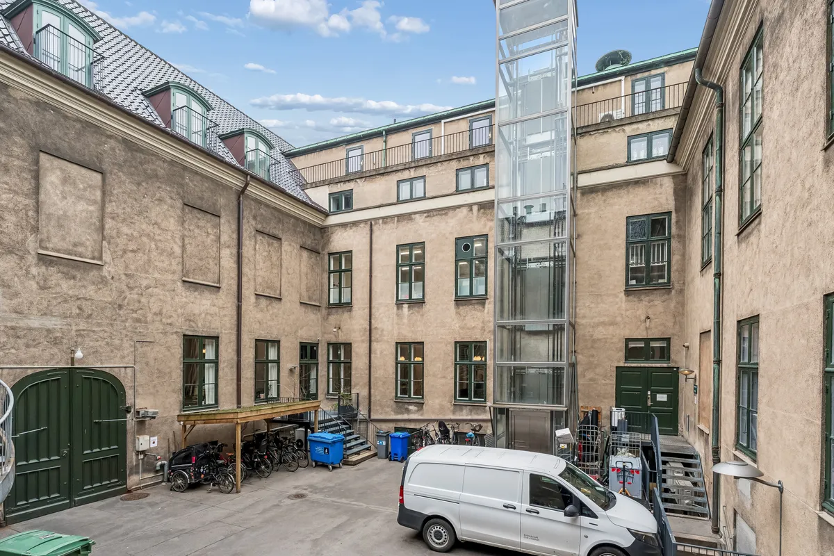 Kontor med parkeringsplads, elevator, cykler og skraldspande i en bygning med lyse vinduer og terrasser