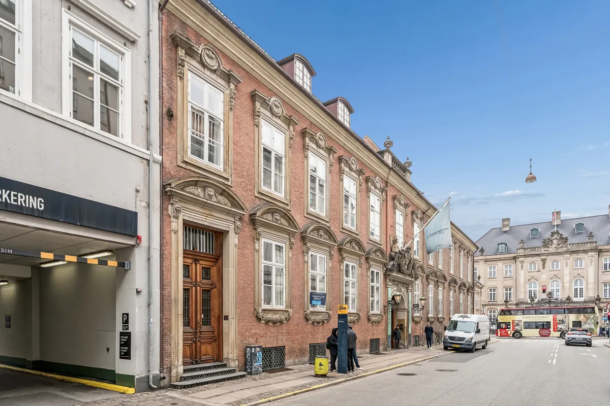 To personer står og venter på bussen i nærheden af et stort rød murstensbygning. Kontor til leje står på en væg. Der er en bil og en bus på vejen