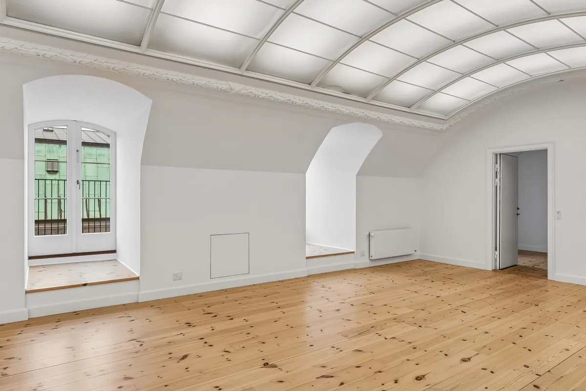 En stor to-rumskontor med højt hælvede loft og lyse trægulve samt et lille spisestueområde med store vinduer og balkon med udsigt til byen