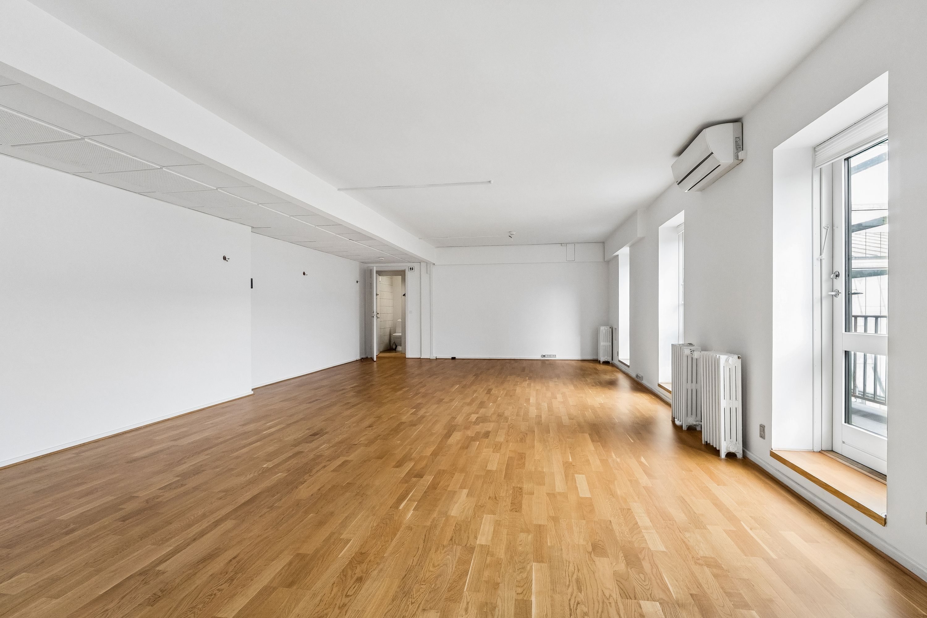 Lukket rum med trægulv og hvide vægge, højt til loftet, en klimaanlæg og radiatorer ved vinduet, glasdøre og hvidt loft, og en åben dør til toilettet