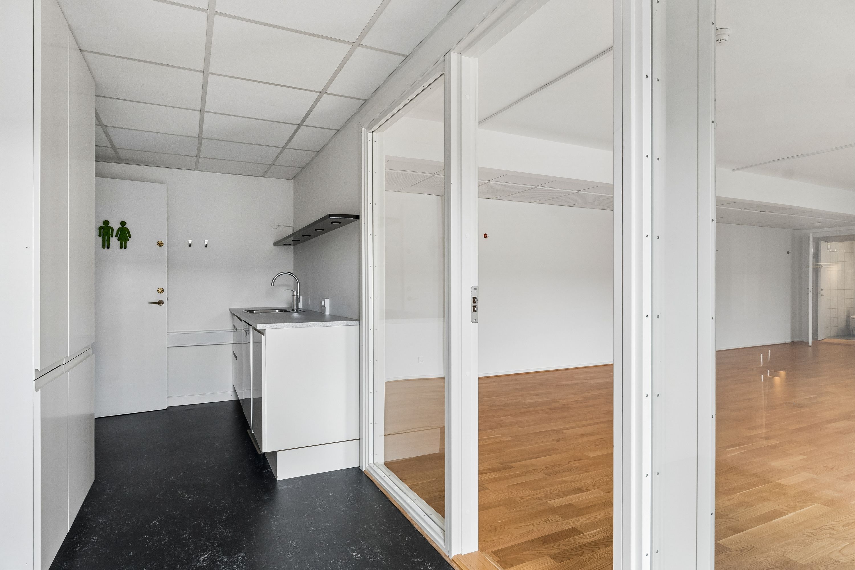 Kontor til leje med 2 store rum, hvidt køkken med lyse trægulve og store vinduer, toiletskilt på væggen, uden møbler og indretning