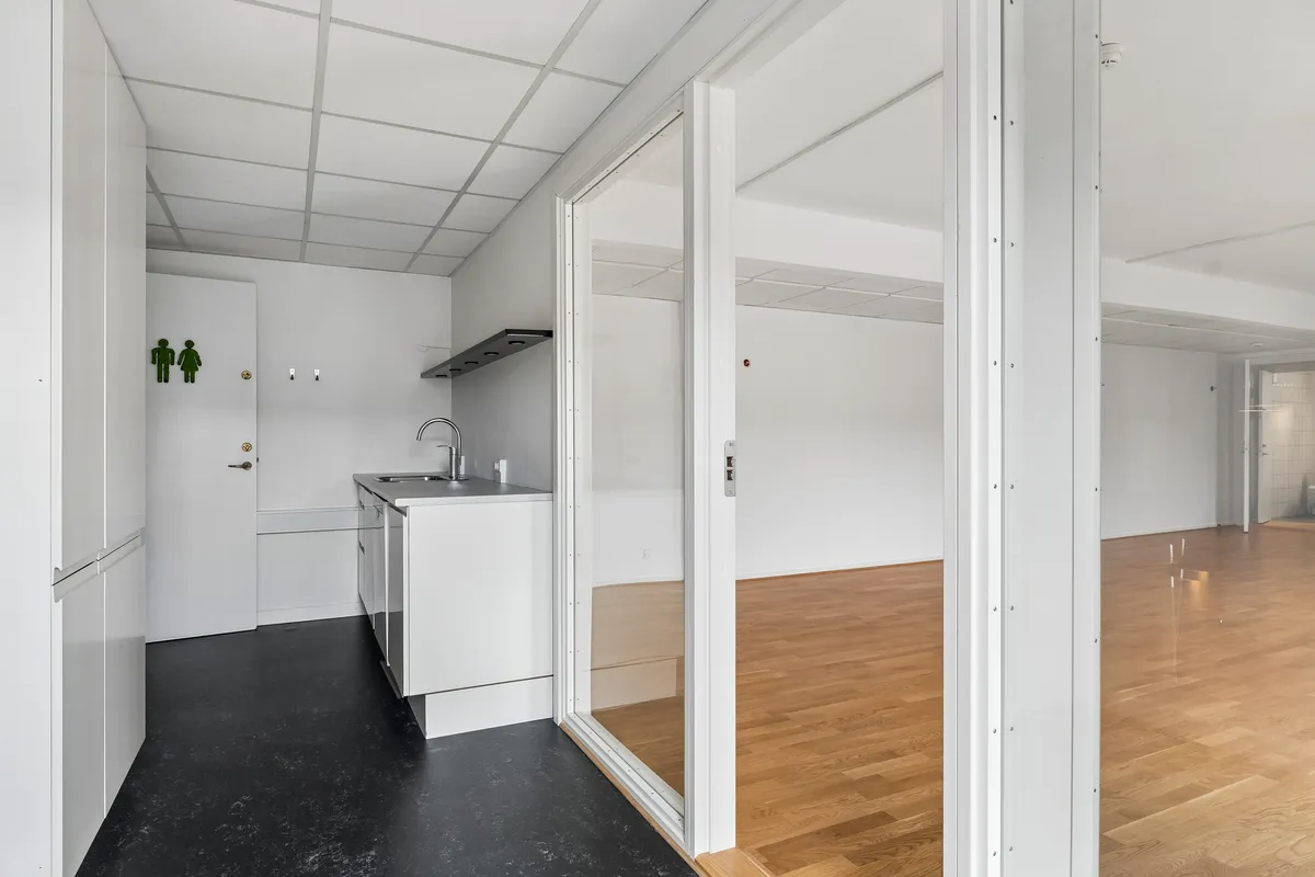 Kontor til leje med 2 store rum, hvidt køkken med lyse trægulve og store vinduer, toiletskilt på væggen, uden møbler og indretning