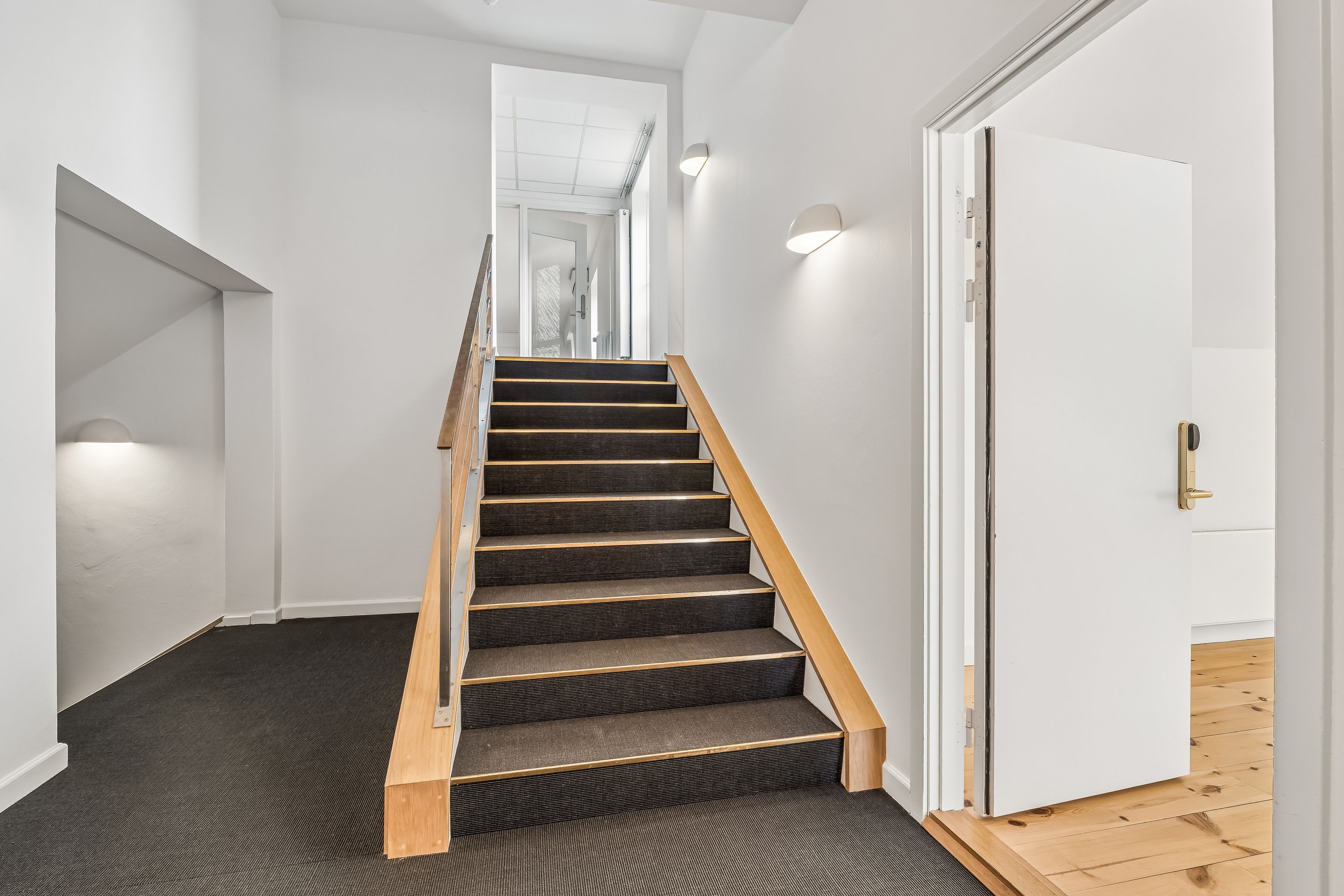 Kontor med lyse trætrapper og hvid væg. Døren til højre er åben med gult dørhåndtag og lys på væggen. På gulvet ligger grå gulvtæppe