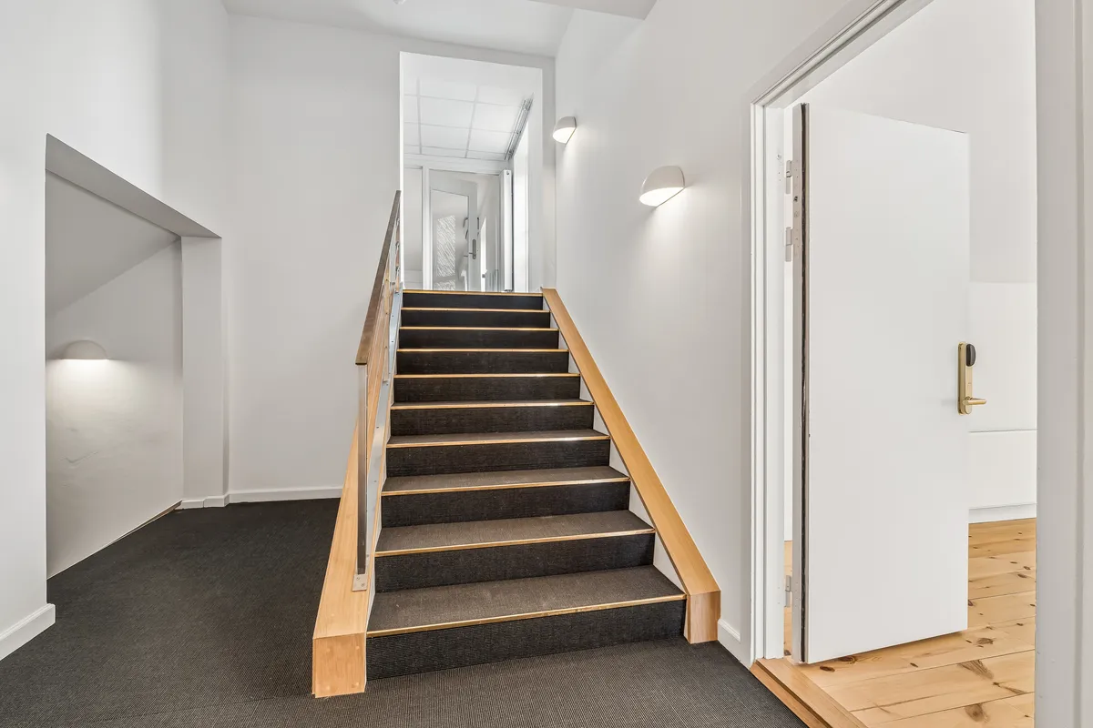 Kontor med lyse trætrapper og hvid væg. Døren til højre er åben med gult dørhåndtag og lys på væggen. På gulvet ligger grå gulvtæppe