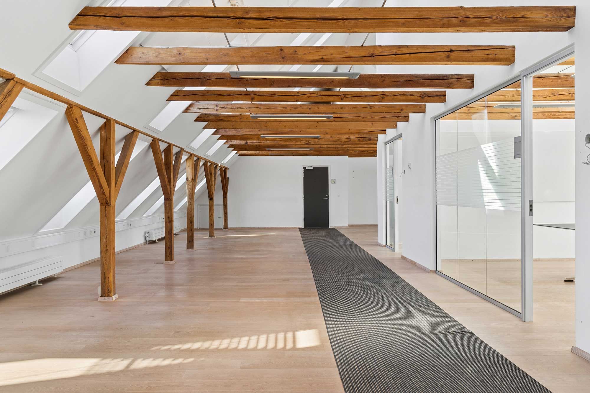 Lyst kontorlokal med lyse loftsbjælke og glasvinduer uden for. Loftsbjælken er lavet af træ og gulvet er lavet af træ. Lokalet er møbleret med skrivebord og stol.